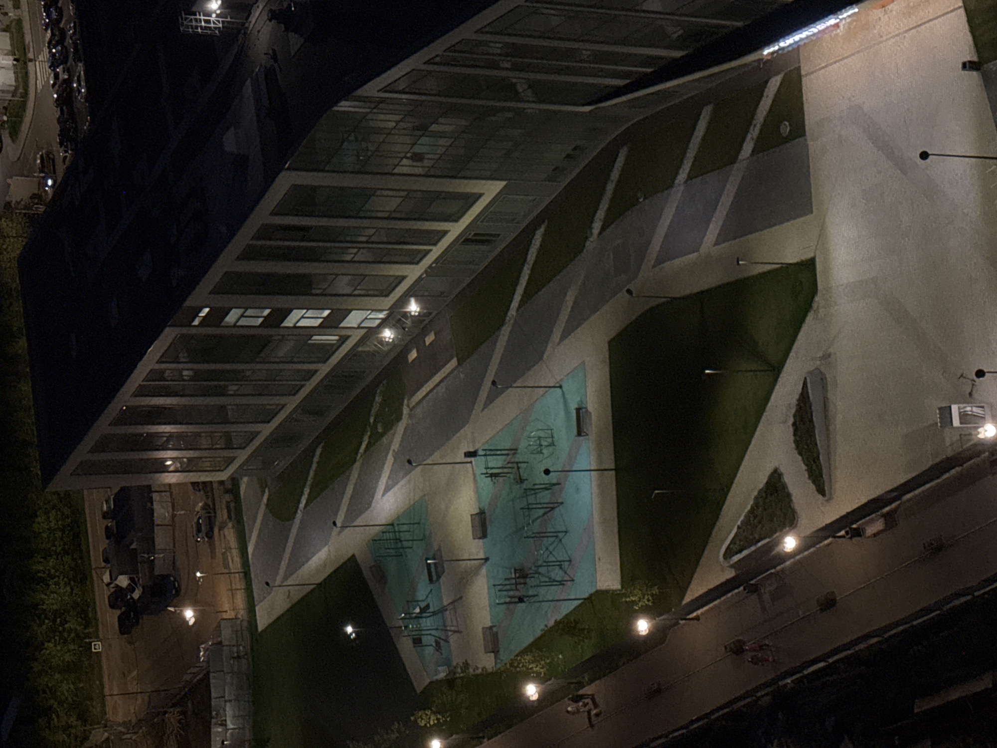 The image shows an aerial view of a park and adjacent buildings in Lyubertsy, Russia at night. A large, modern building with a glass roof and rectangular windows occupies the upper left portion of the frame. A park area is located beside the building, featuring a walkway with geometric patterns of light and dark pavement and grass. There is an outdoor exercise area with metallic equipment visible. A road borders the park, with pedestrians walking along it. Streetlights illuminate the walkway and the road. Vehicles are parked near the building, including what seems to be a large moving truck.