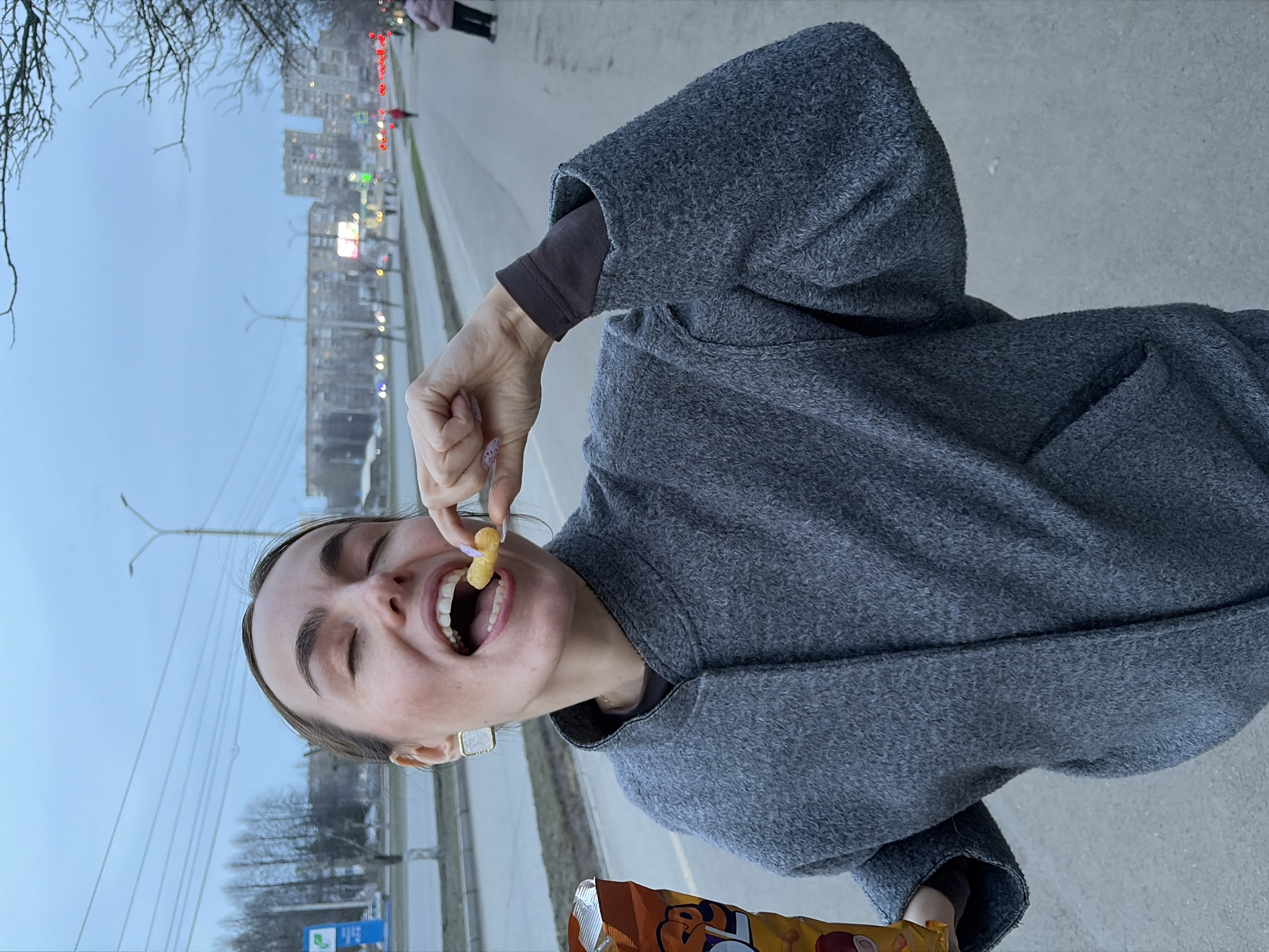 A woman with light skin and brown hair pulled back is eating a cheese puff snack in an outdoor setting. She is wearing a gray coat over a brown long-sleeved shirt and a square earring. She holds a bag of cheese puffs in her left hand. The background shows an urban landscape, including buildings, street lights, and a road with a pedestrian. Based on the urban landscape featuring building and street lamps and the blue-green sign with white text, it's likely the location is a road in the city of Izhevsk, Russia.