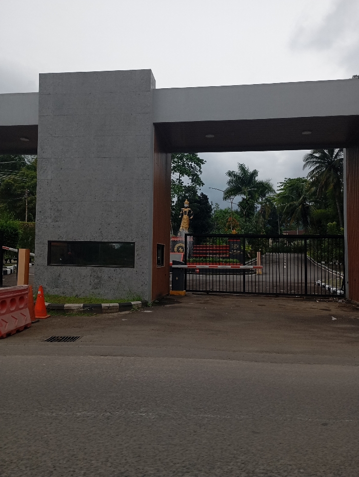 The image shows a gated entrance to a property in Kecamatan Kutawaringin, Indonesia. The gate is made of black metal bars and is closed. There is a guard booth on the left side of the gate. The booth is made of concrete and has a small window. The booth is topped with a gray roof. There is a red and white traffic cone on the left side of the guard booth. The gate is under a large, white, concrete archway with a brown wood accent on the top. The gate is flanked by two concrete pillars. The gate is covered in red and black paint. Behind the gate, a paved road leads to a small statue of a man in the middle distance. The statue is located on the left side of the road. The man is wearing a gold-colored outfit. On the right side of the road, there is a green area with trees. The sky is gray and cloudy. The road leading to the gate is paved and appears to be gray asphalt.
