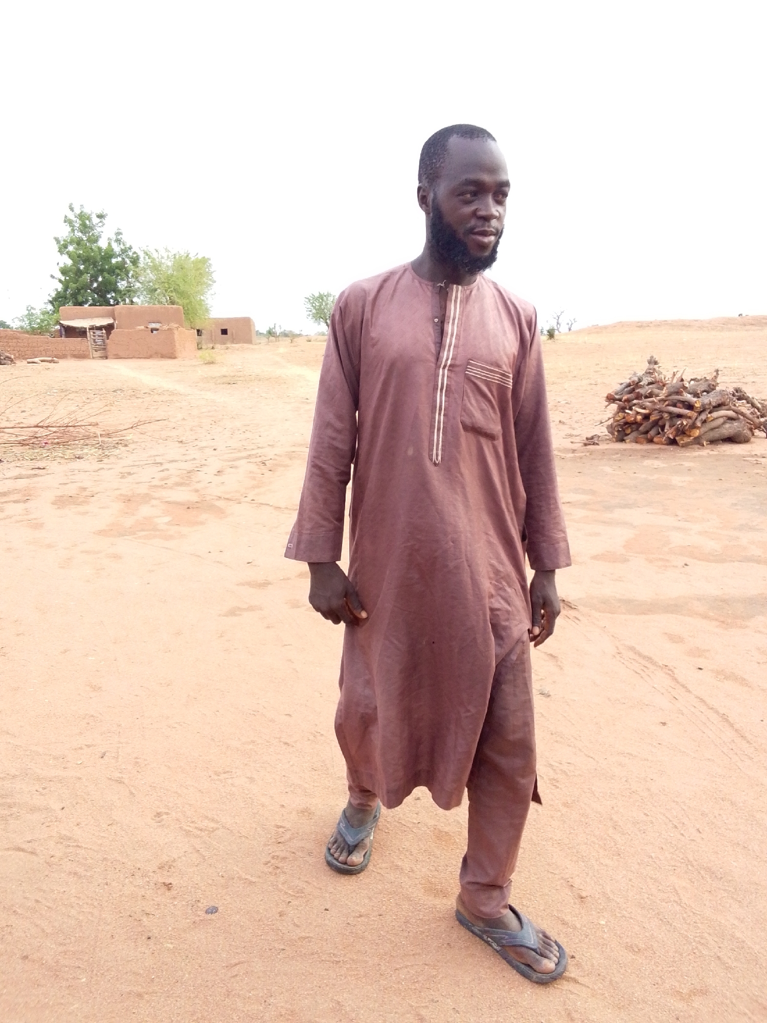 A man stands in a sandy, arid environment in Mulke Barikin Sayejo, Nigeria. He wears a long, brown robe-like garment and simple sandals. Behind him are low, mud-brick buildings and a pile of cut wood. The sky is overcast.