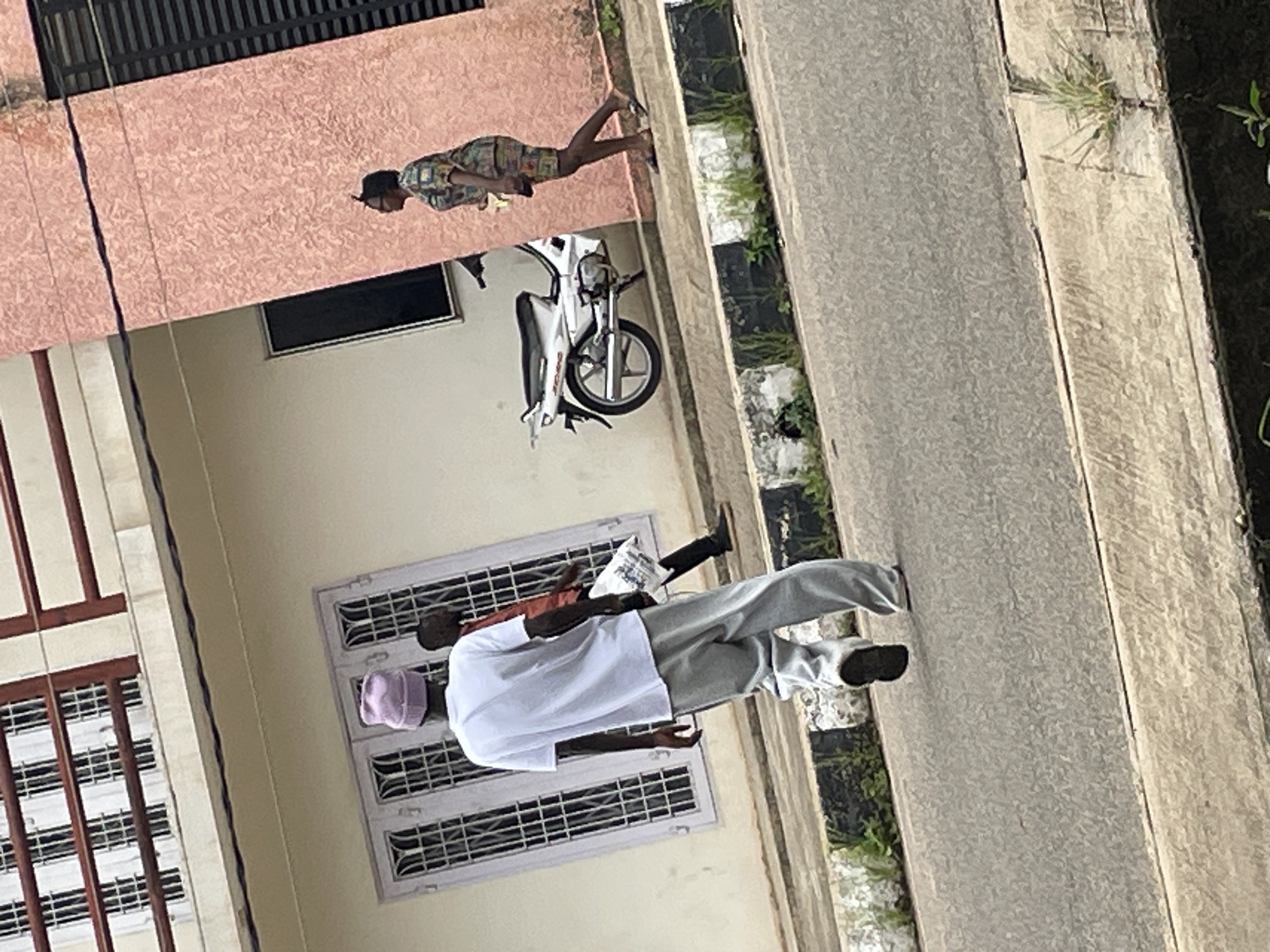 The image depicts a street scene in Jos, Nigeria, with a focus on pedestrians and a motorcycle. In the foreground, a person wearing a purple beanie and gray sweatpants walks away from the camera, their back to the viewer. To their right, another person in an orange shirt is partially visible, carrying a white bag. In the background, a white motorcycle is parked. Further to the right, a woman wearing a patterned dress and skirt walks away from the motorcycle. The environment includes a building with white and pink stucco walls and barred windows. The street has a concrete curb with grass growing along its edges.