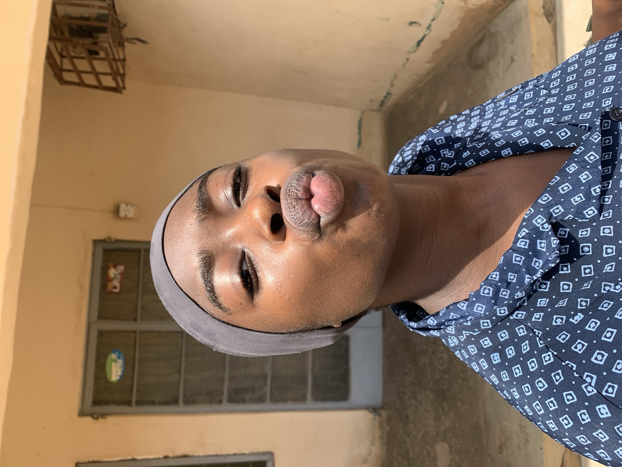 A person with brown skin, wearing a gray head covering and a blue patterned shirt, is making a kissing face. The background includes a yellow wall with a barred window and a caged structure near the ceiling. The scene appears to be outdoors with a rough wall behind the person. The location is Rafin Par, Nigeria.