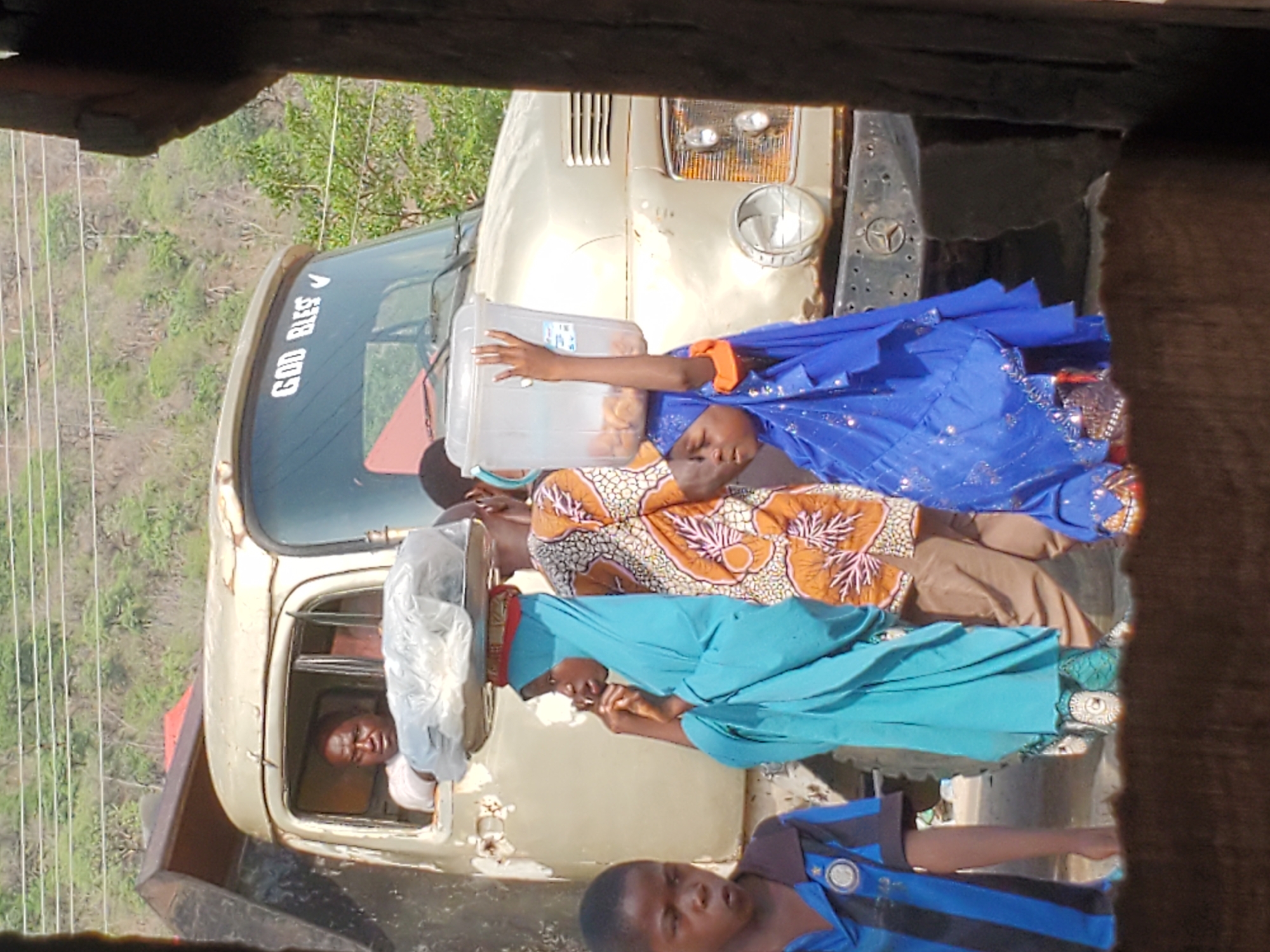 In Mararaba, Nigeria, a medium, eye-level shot shows four children riding on the hood of an old, beige truck. The truck, displaying the phrase "GOD BLESS" on its windshield, is parked in an outdoor environment with trees visible in the background. The driver, an adult male, sits in the driver’s seat of the vehicle. The first child is positioned closest to the front of the truck, wearing a blue and black striped shirt. The second child wears a light blue outfit. The third is attired in an orange patterned outfit. The fourth, closest to the windshield, wears a sparkly blue outfit and holds a translucent container.