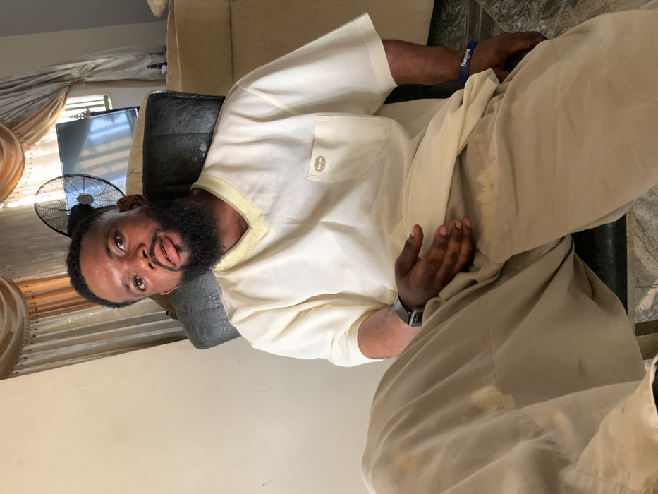 The scene is situated within Eika-Adagu New Layout, Nigeria. An adult male is seated indoors, positioned centrally and facing the viewer directly. He has a dark, well-groomed beard and mustache, with short, dark hair. He wears a light cream-colored, short-sleeved V-neck top featuring a small pocket with a light brown label on the left chest. His lower garment consists of light brown or khaki trousers, displaying discolouration or stains on the left thigh and groin area. A dark-colored smartwatch with a black strap is on his left wrist, and a blue wristband with partial white text is on his right wrist. His left hand rests open on his left thigh. The man is seated in a dark-colored chair or sofa. In the background, a dark standing fan with an open cage is visible to the right of the man's head, partially obscuring a wall-mounted flat-screen television. Cream-colored curtains with decorative valances are present in the background. A light-colored wall is to the left of the subject, and tiled flooring, possibly marble or polished stone, is visible at the bottom of the frame. A portion of another light-colored sofa is discernible in the far right background.