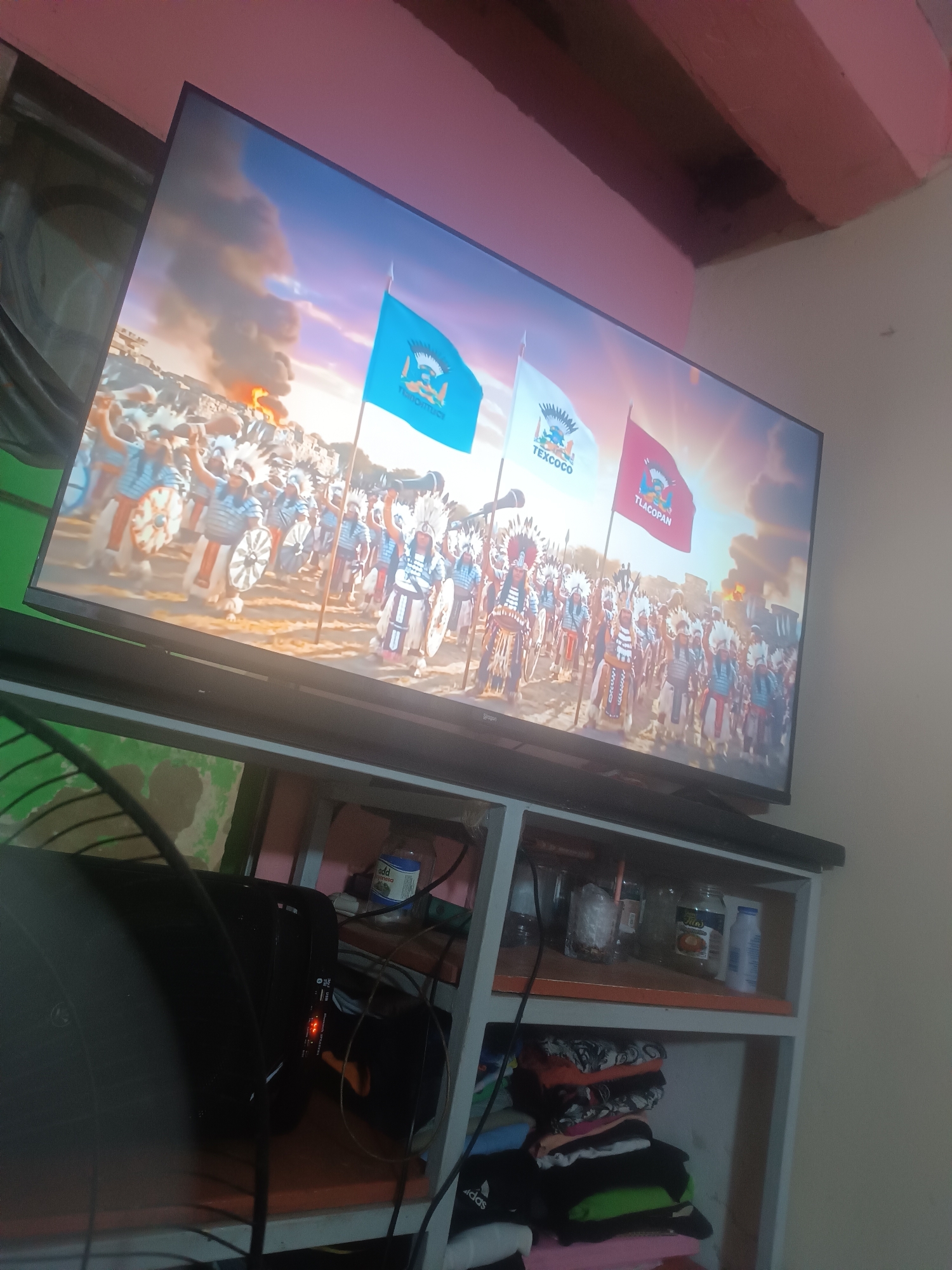 The image shows a TV displaying a scene with indigenous warriors carrying flags labeled "Texcoco" and "Tlacopan". The setting appears to be historical. The TV is placed on a white and orange shelf unit. The lower shelves hold items including a black computer tower, jars, a bottle, and folded clothes. The room has pink accents on the ceiling, with light yellow walls. The location and city of Porlamar, Venezuela, cannot be determined from the image.