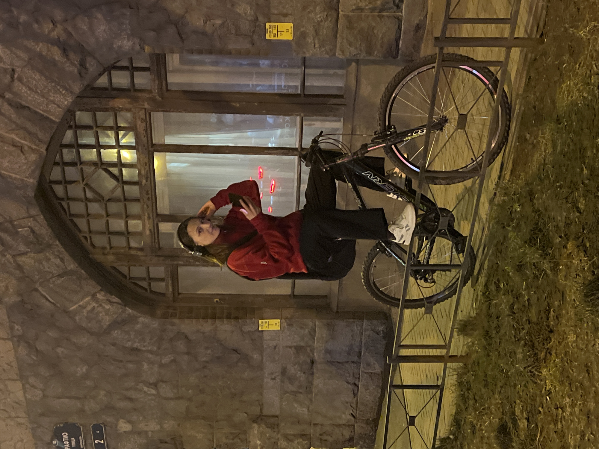 This forensic description details an evening urban scene in Saint Petersburg, Russia. A young adult female is prominently featured, seated on a black Stern mountain bicycle that is balanced on a low, dark metal fence. She has dark hair, wears a red long-sleeved top, black trousers, white athletic shoes, and silver over-ear headphones. She holds a dark smartphone in her right hand, her left hand near her headphone, and looks directly at the camera with a subtle smile. A dark backpack is visible on her back.
Behind the female and bicycle, a rugged stone-faced building with a large, arched, multi-paned window forms the backdrop. The window shows reflections and internal illumination, including several horizontal red light streaks. To the left of the window, on the stone wall, a blue street sign is partially visible, displaying "РАФТИО УЛИЦА" and the number "2". A small yellow utility sign is also affixed to the wall. The ground consists of a paved sidewalk immediately behind the fence and a strip of sparse grass and dirt in front of it. The entire scene is illuminated by artificial, warm-toned street lighting, indicating nighttime conditions.