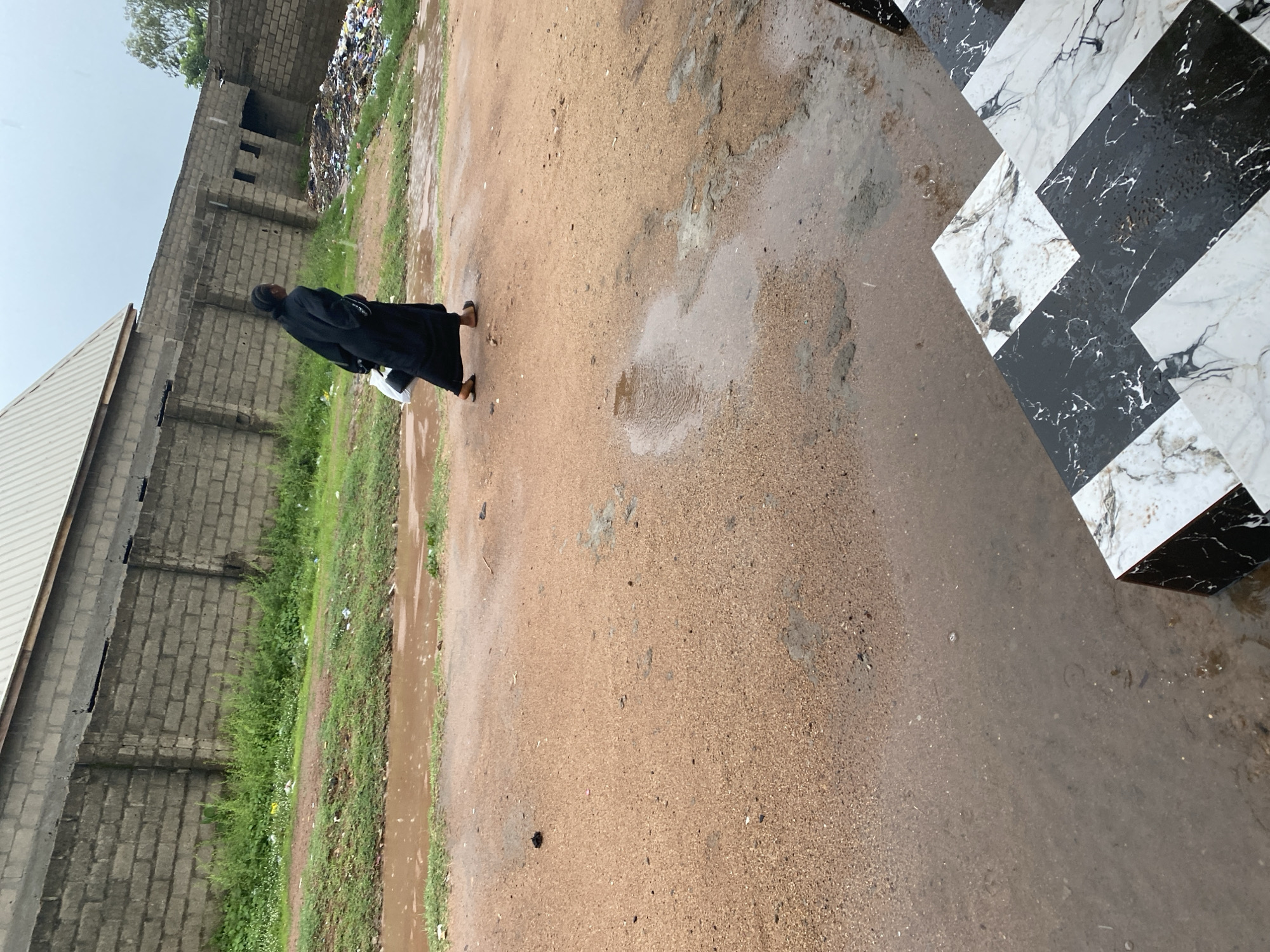 A lone figure walks on a dirt path in Jalingo, Nigeria, with a brick wall and a building visible in the background. The path is wet, with several puddles reflecting the overcast sky. To the right of the path, there is a pile of garbage. In the foreground, a set of stairs with a black and white marble pattern is partially visible. The person is wearing a dark, flowing garment and carrying a white bag in their left hand. The person is walking away from the camera, towards the right of the frame. The overall scene suggests an outdoor setting, possibly after rainfall, in an area that appears to be under development or with some unkempt areas.