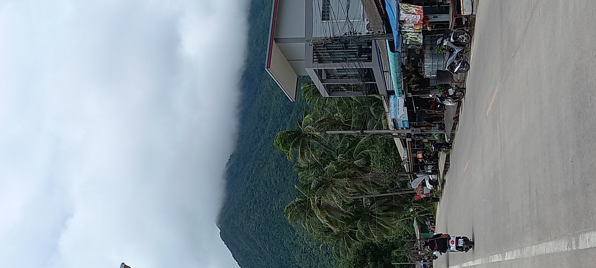 The image captures a street scene in ตำบลเกาะพะงัน, Thailand. A concrete road stretches into the distance, partially obscured by a motorcycle with two riders. The roadside features parked motorcycles and local businesses under a multi-story building. Tropical palm trees line the street, beyond which lies a lush, green mountain shrouded in low-hanging clouds. The weather appears overcast. Overhead, electrical wires are visible, crisscrossing the scene.