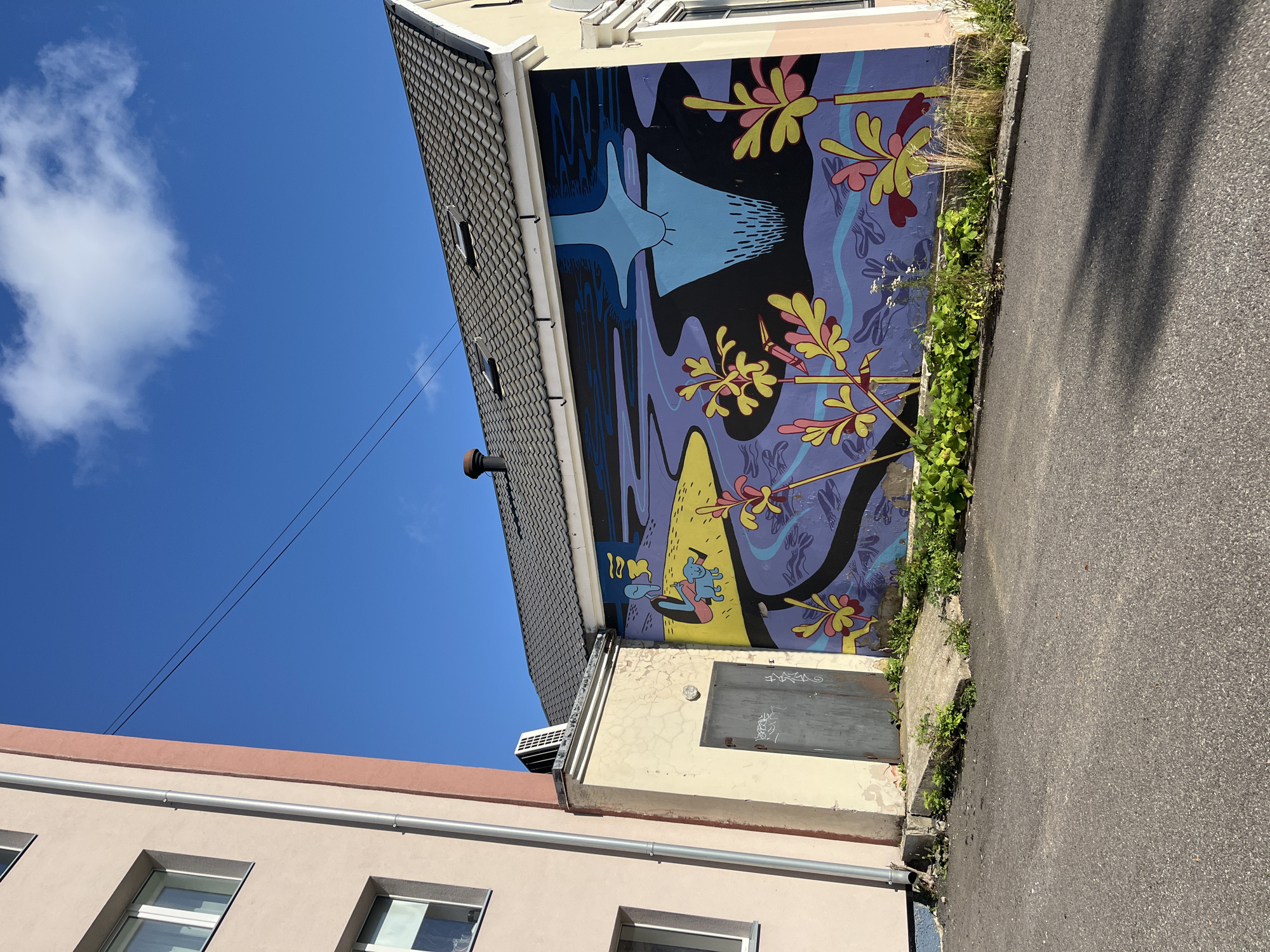 The image shows a mural on the side of a building in Kohtla-Järve linn, Estonia. The mural depicts a stylized scene with a blue figure sitting by a yellow body of water, with a blue abstract shape reflected in the water. There are also yellow and pink plant-like elements in the foreground and scattered across the mural.
To the left of the mural is a light-colored building with multiple windows. A drainpipe runs vertically along this building, and an air conditioning unit is visible on the roof of a smaller attached structure. This structure has a weathered, light-colored wall with cracks and a gray metal door with graffiti.
The mural covers a significant portion of the wall of a building with a dark, tiled roof. The sky above is bright blue with a few white clouds. The ground in front of the mural is asphalt, with some patches of grass and weeds growing along the base of the wall and on a concrete ramp leading to the gray door. The scene is illuminated by sunlight, casting shadows on the asphalt.