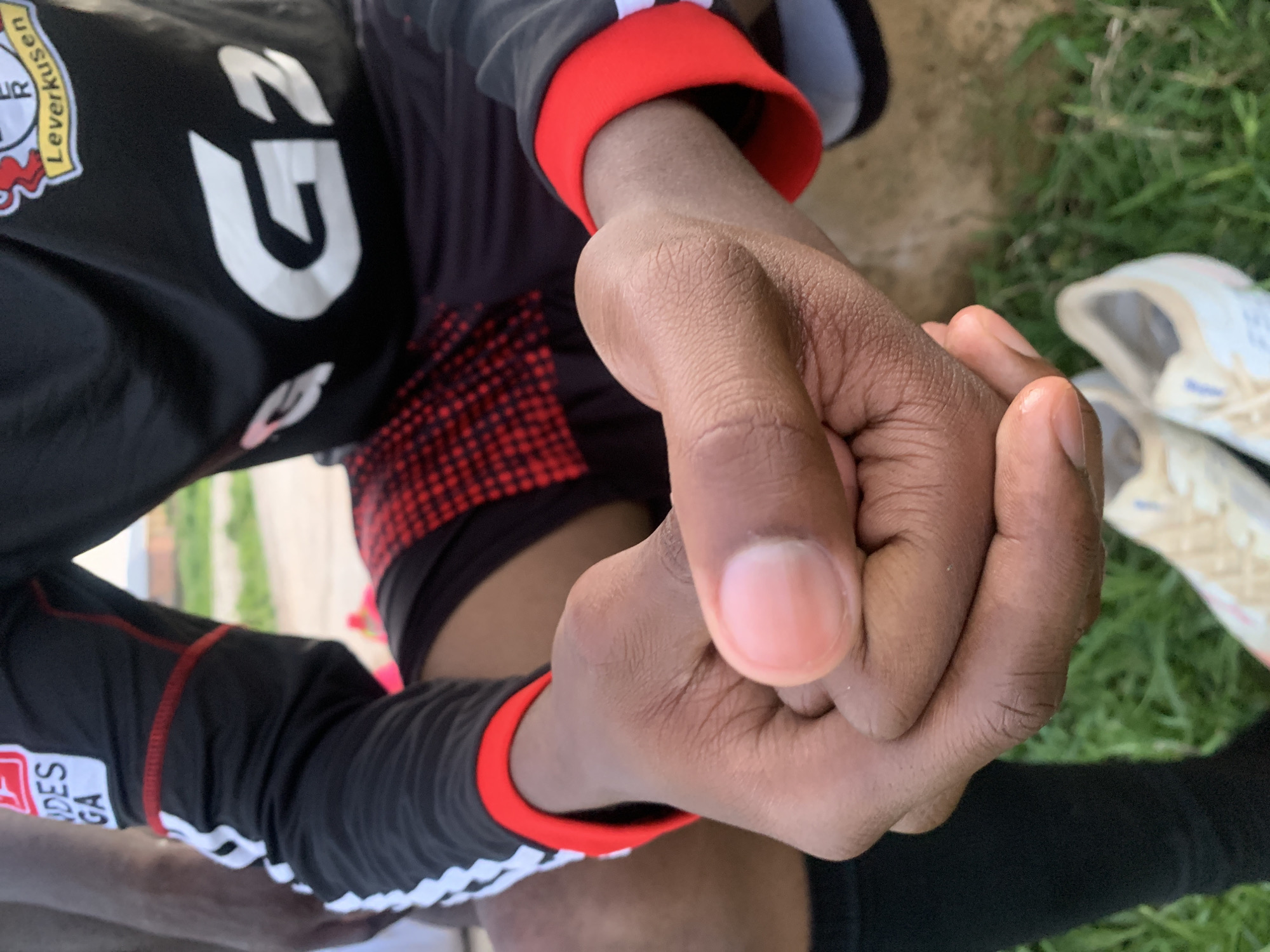 The image shows a close-up view of a person's hands, one fist clenched and the other partially open, against a backdrop of green grass and some dirt. The person is wearing a black sports jersey with red accents on the sleeves and collar. Visible text on the jersey includes "Leverkusen" and what appears to be the number "23" in large white font. There is also a red and black checkered pattern on the fabric. The lighting suggests it could be daytime. The setting appears to be outdoors, possibly a sports field, given the attire and the grass. The focus is on the hands and the texture of the skin, with some of the sporting apparel visible. The crops provide closer views of the jersey details and the hand's knuckles and fingernails.