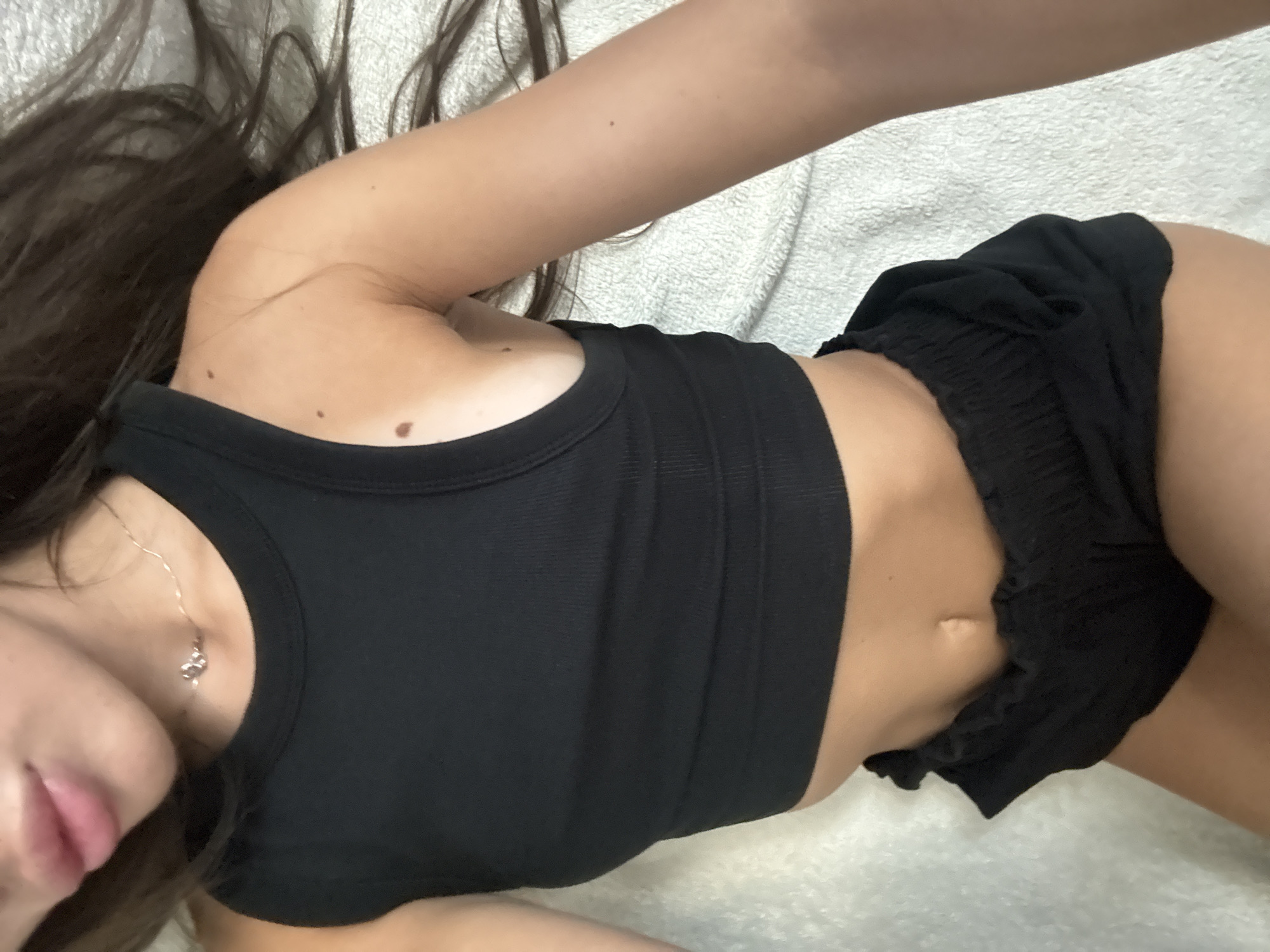 This is a close-up, overhead shot of a person lying on a textured, white surface. The person has long, dark hair that extends across the frame. They are wearing a black tank top and black shorts. A delicate silver necklace with a small pendant is visible around their neck. The lighting is soft, suggesting an indoor setting. The person's skin tone is fair with visible moles on their shoulder and arm. The overall mood is relaxed and casual. There is no visible text in the image.