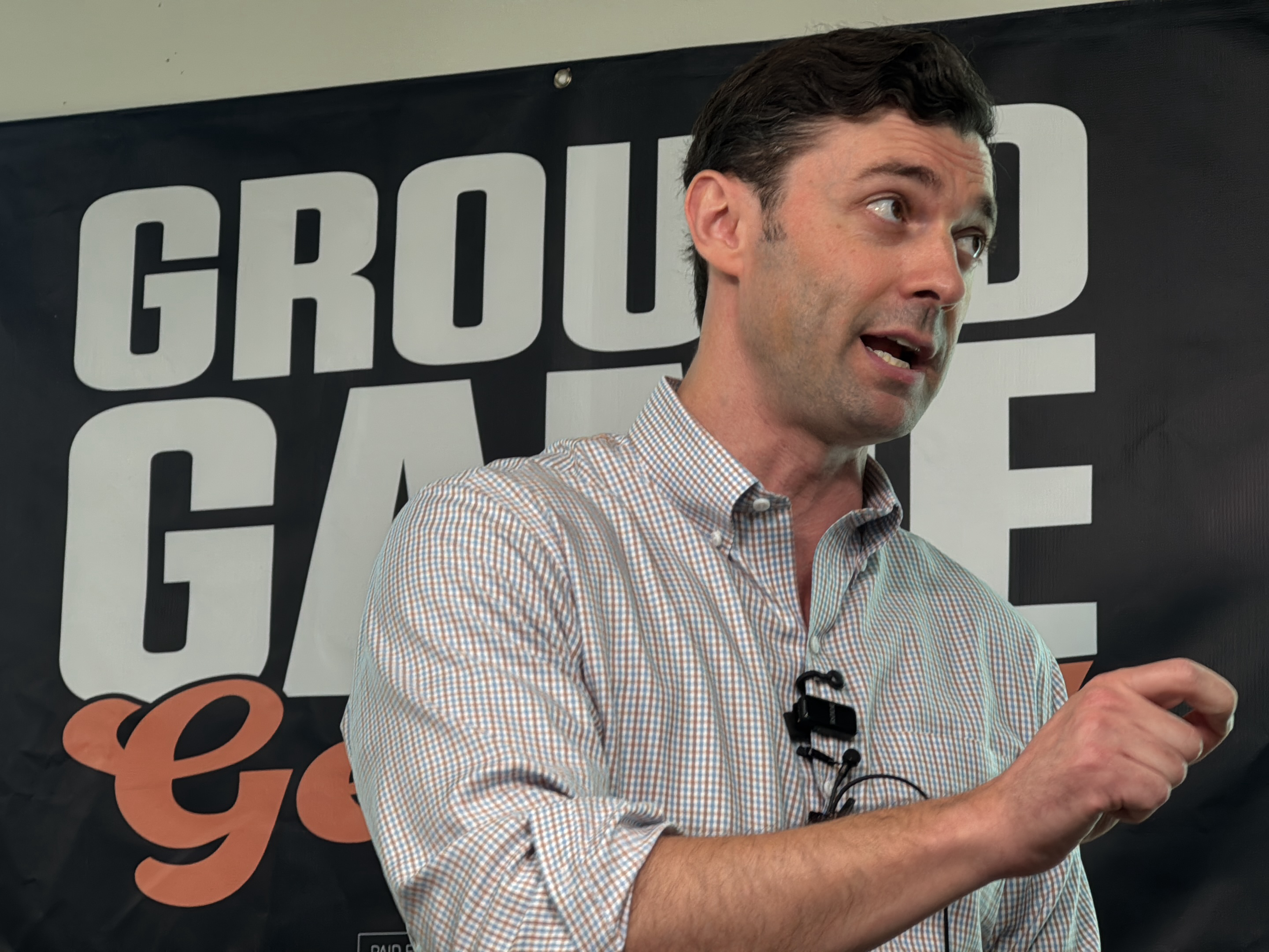 The image shows a man standing in front of a black banner with the words "GROUND GAME" printed in white and orange letters. The man is wearing a light-colored button-down shirt with a small microphone clipped to it. He is gesturing with his right hand, his mouth slightly open as if speaking. The location is likely Decatur, United States, based on the content of the banner.