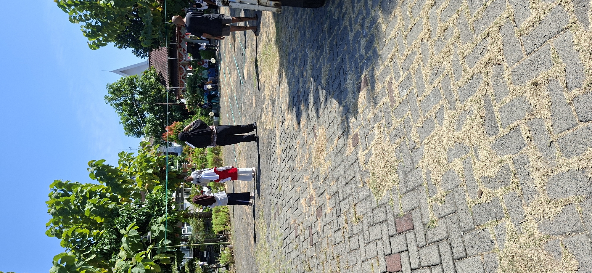 The image depicts an outdoor scene, possibly a public space or a park, on a clear day. In the foreground, a paved area with interlocking bricks is visible, interspersed with patches of dry grass and straw. Several people are present in the midground and background.
In the midground, three individuals are standing in what appears to be an informal gathering or activity. Two of them are young females wearing hijabs, one with a brown hijab and the other in a white hijab with a red top. They are facing away from the camera, towards a man in a grey shirt who is also facing them. Further back, another person in a black hijab and black pants is facing away from the camera, with their right hand near their head. To the right, a man wearing a black t-shirt and shorts is standing and facing the left.
In the background, there is a building with a tiled roof and a white pointed structure, possibly a church steeple or a similar architectural feature. Trees with lush green foliage are scattered throughout the scene, framing the view of the sky and the building. Some decorative elements, possibly flags or banners, are hanging from wires strung between poles. A group of people are seated in the background, observing an activity. The environment suggests a sunny day with clear blue skies.
The exact location in the city of Surabaya, Indonesia cannot be definitively determined from the visual information alone. However, the presence of specific architectural styles, vegetation, and the attire of some individuals might suggest a location within Indonesia. Without further context or identifiable landmarks, pinpointing the precise spot in Surabaya is not possible.