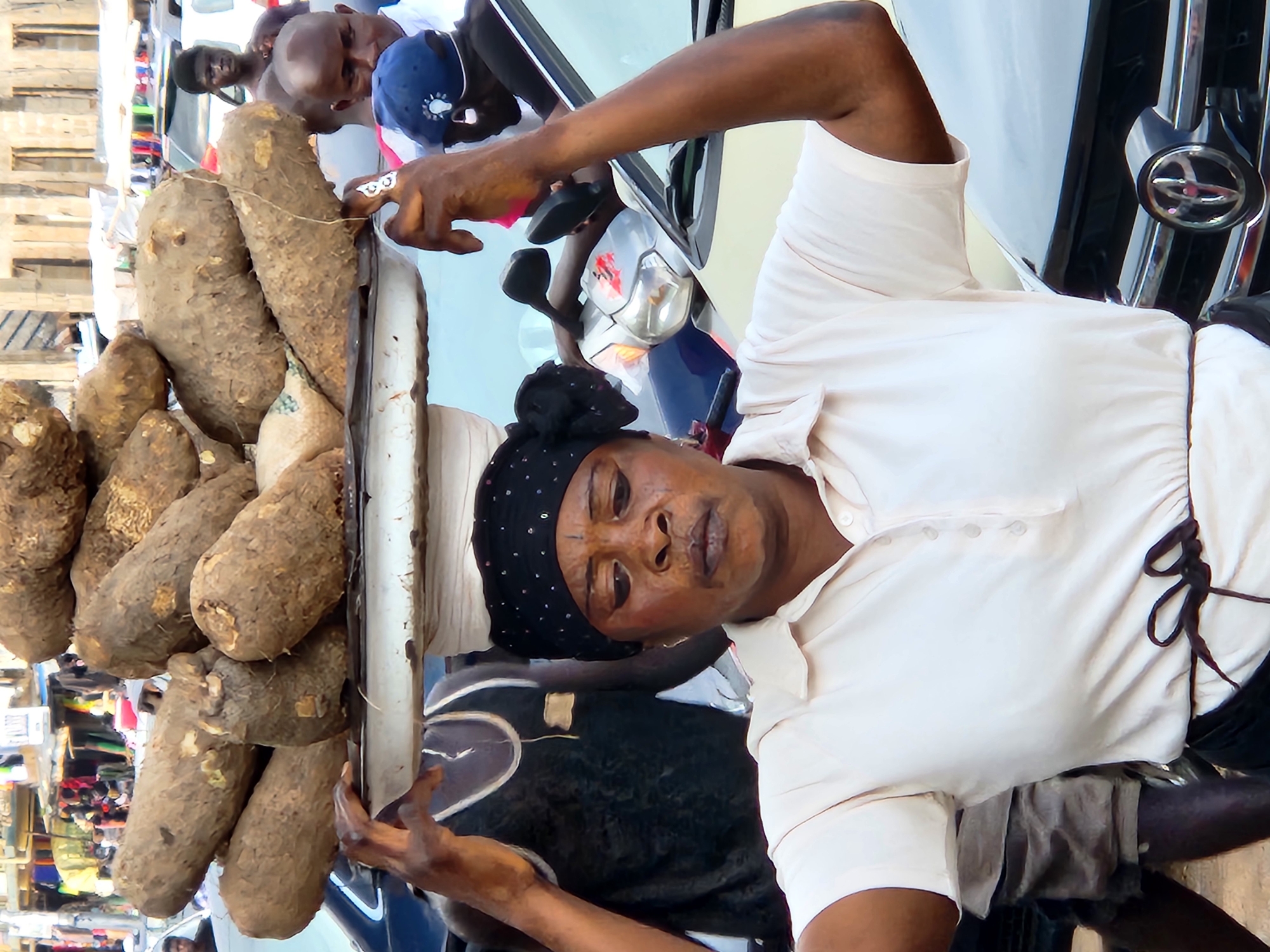 A woman wearing a light-colored polo shirt and a black headscarf with white polka dots balances a large tray of yams on her head. She is in a bustling outdoor market in Jos, Nigeria. In the foreground, a black car is partially visible. Behind the woman, several other people are seen, including a man in a black tank top to her left and a man in a blue baseball cap to her right. The background shows market stalls with various goods and more people. The overall scene depicts a vibrant and busy marketplace.