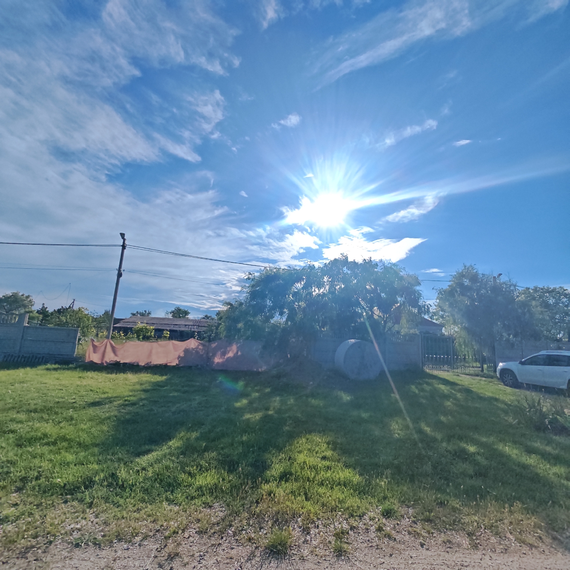 The photograph shows a sunny day in an outdoor setting, likely a residential area in Unknown. The sky is bright blue with scattered white clouds and the sun high above, creating a strong glare. Below the sky, there is a tree casting a shadow on a grassy lawn. A grey fence runs along the property line, with what appears to be an orange sheet or fabric hanging on one section. In the background, there is a glimpse of houses and a utility pole with wires. A white car is parked to the right of the fence. The foreground shows a gravel or dirt path leading to the lawn.
