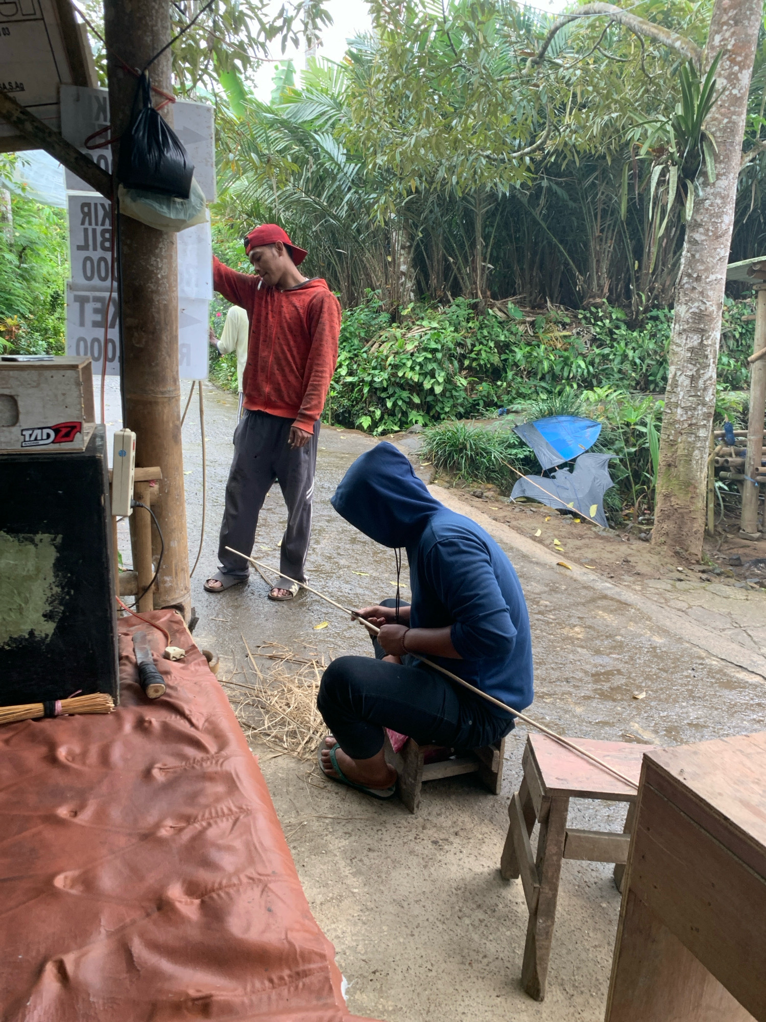 Two individuals are engaged in crafting kites. One person, wearing a red hooded sweatshirt and a red baseball cap, stands near a tree with a black bag hanging from it. They are looking down and appear to be smoking a cigarette. The other person, wearing a blue hooded sweatshirt, is seated on a small wooden stool, intently working on a kite. They are holding thin wooden sticks, which are likely part of the kite's frame, in their hands.
The scene is set outdoors, with lush green foliage and trees forming a dense backdrop. The ground is wet, suggesting recent rain. In the background, two partially assembled kites, one blue and the other gray, are visible, lying on the ground amidst the vegetation. There's a wooden structure to the left of the image with some boxes and equipment, including what looks like a speaker. To the right, another wooden structure, possibly a table or stand, is present. The overall impression is of an outdoor workshop or a casual gathering where kite-making is taking place. The location is identified as Pronojiwo, Indonesia.