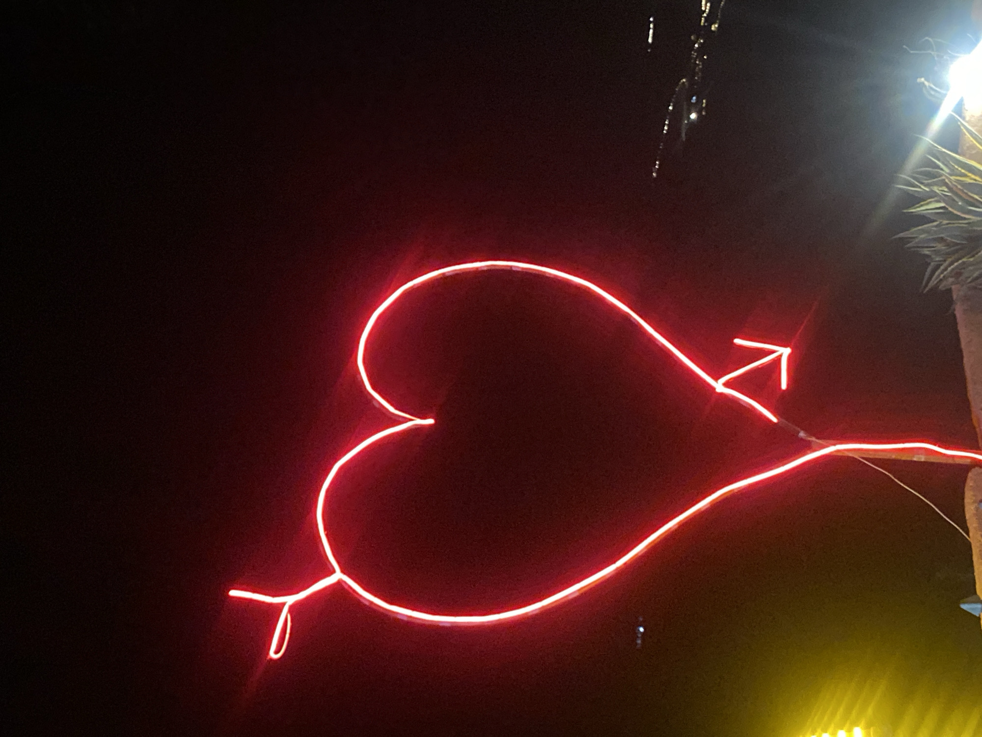 A neon red heart-shaped sign with an arrow piercing it, illuminated against a dark night sky. The sign is positioned in the foreground. In the middle ground, there are some blurry lights and structures in the distance, possibly indicating a settlement along a coastline. In the bottom right corner, a plant with spiky leaves is visible, along with some bright light flares. A portion of a yellow light source is visible in the lower left. The scene is set outdoors, likely in a location with a view of a distant, illuminated area. The image appears to have been taken in Agafay, Morocco.