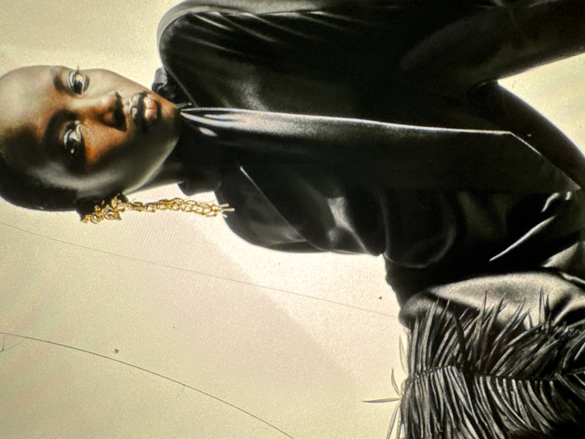 A close-up shot features a model with dark skin and short hair, adorned with a large, intricate gold earring that dangles from her left ear. She is wearing a high-necked, black garment that appears to be made of a smooth, reflective fabric. The lower left of the image reveals a textured detail on her clothing, possibly ruffles or pleats, rendered in a dark, matte material. The background is a plain, light-colored surface with subtle lines and faint marks, suggesting an indoor studio setting or a wall. The lighting creates strong highlights and shadows, emphasizing the model's features and the textures of her attire. There is no indication of the city of Protasovo, Russia, in this image.