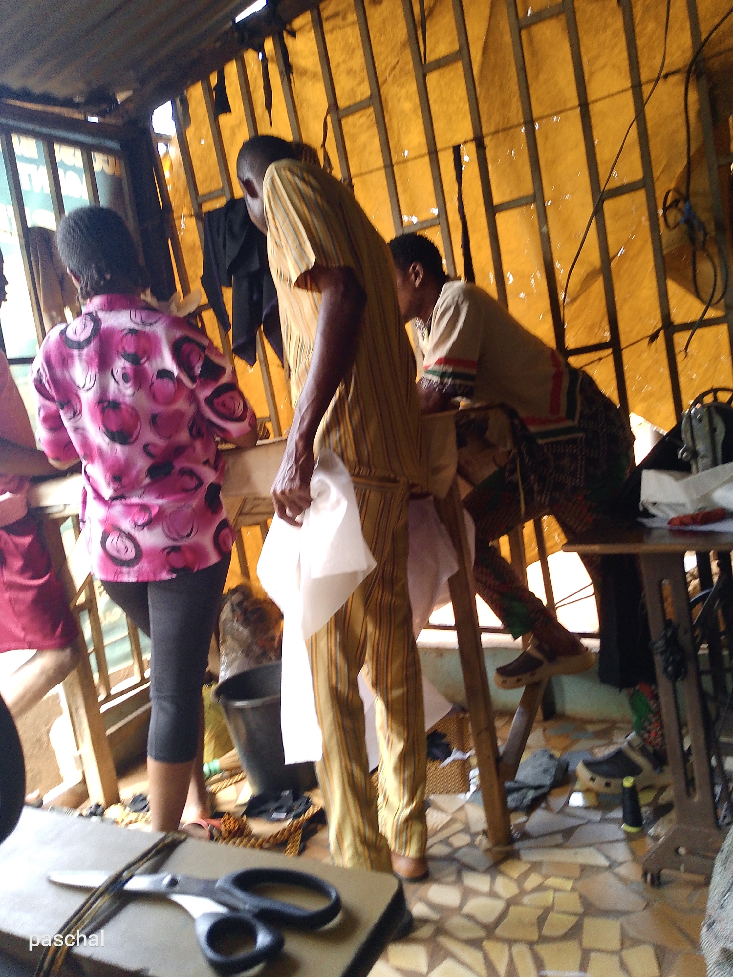The photograph shows the interior of a tailoring shop in Nsukka, Nigeria. Three people are visible. One woman wearing a pink and black patterned shirt and black leggings stands near a work table. Another man in a striped yellow suit is holding fabric. Another man wearing a shirt with green and red details is leaning over a sewing machine. The walls of the shop appear to be made of wood and yellow fabric. A metal bucket sits on the floor, and a pair of scissors are on a work table in the foreground. The floor is tiled in a mosaic pattern.