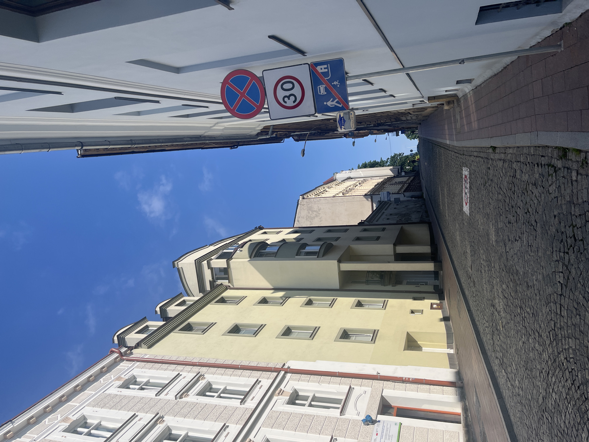 A sunlit, narrow, cobblestone street slopes uphill in an urban setting, lined by multi-story buildings. On the left, buildings with light gray and pale yellow facades feature visible windows and architectural details. On the right, a white building dominates the foreground, with a metal pole mounted with three traffic signs: a red circle with a blue cross indicating no stopping, a square sign with "30" in a red circle indicating a speed limit of 30, and a rectangular blue sign showing a car and pedestrian with a diagonal line through them, signifying no entry for vehicles and pedestrians. The sky above is a clear, vibrant blue with a few faint clouds. A brick sidewalk runs alongside the white building on the right.