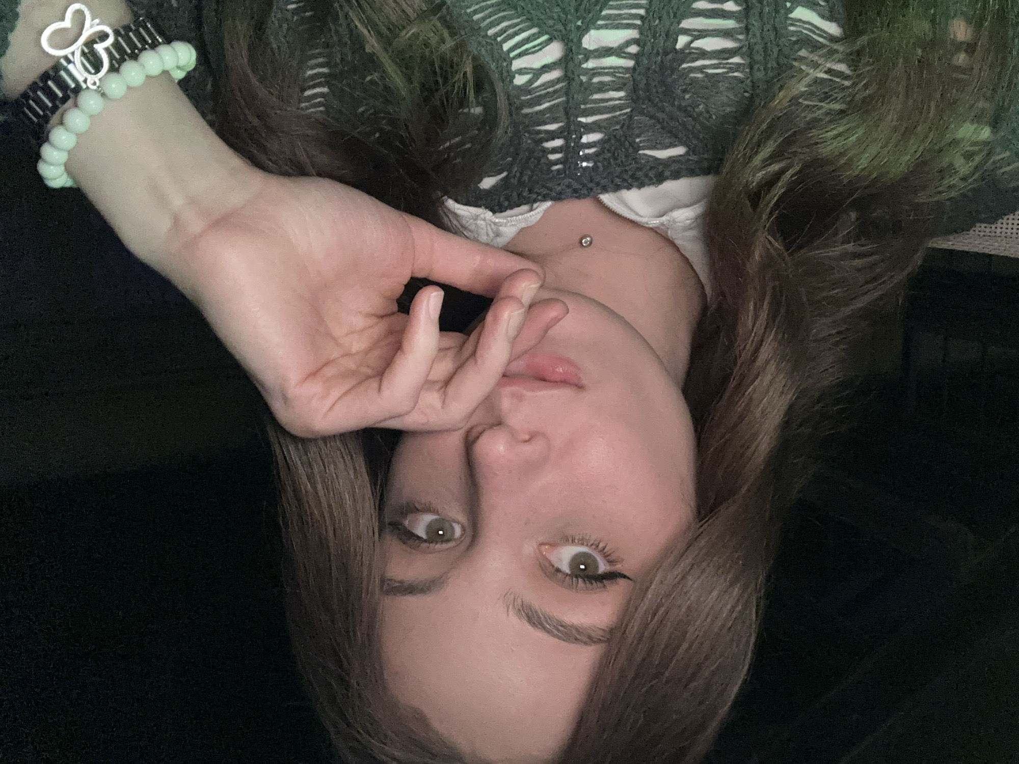 A young woman lies in an unknown location, looking into the camera. She wears a gray, knit sweater with a white shirt beneath. She has a silver-colored piercing on her upper chest. She wears two bracelets on her wrist: a black metallic one and a light-green beaded one with a heart charm. Her hair is brown and partially covers her face. The background is a dark, indistinct color. No other location information is discernable from the image.
