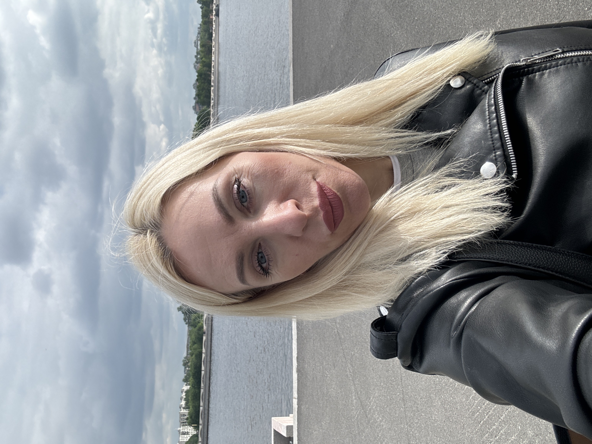 A fair-skinned woman with blonde hair is the primary subject of this portrait-style photograph. Her hair is partially framing her face and draped over a black leather jacket she is wearing. The jacket is zipped and features silver buttons.
The woman has blue eyes and is wearing makeup, including lipstick. Her gaze is directed towards the camera.
The background consists of a body of water, presumably a river or lake, with buildings visible along the shoreline in the distance. The sky above is cloudy. A concrete walkway or promenade is visible in the foreground.
Based on the information given in the prompt, the location is in the city of Unknown, Unknown.
