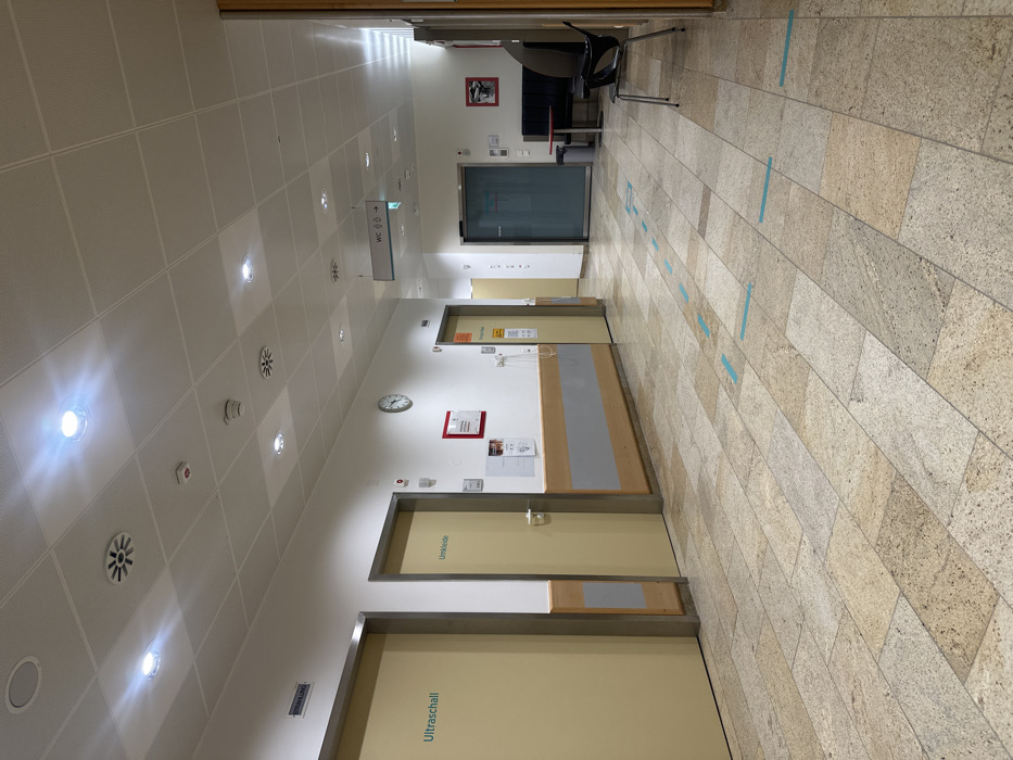 An indoor, slightly tilted shot depicts a hospital hallway with tiled flooring and a suspended ceiling featuring round vents and recessed lighting. On one side, doors labeled "Ultraschall" and "Umkleide" suggest medical facilities. A reception desk stands along the wall. Further down, there's an elevator. Blue tape markings are spaced intermittently on the floor. A chair and desk sit at the end of the hallway.