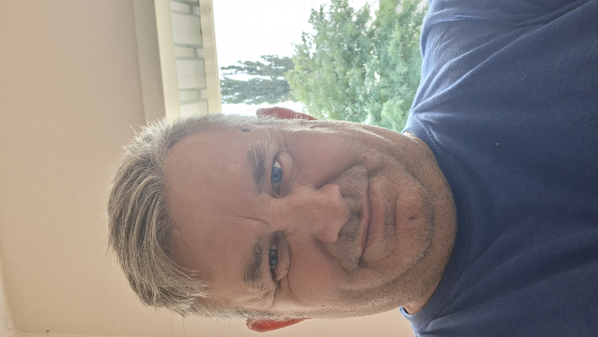 A middle-aged man with grey hair and blue eyes is captured in a selfie-style portrait. He is wearing a blue t-shirt and has a fair complexion. His facial expression is neutral, and he is looking directly at the camera. The background appears to be a window with greenery and trees visible outside, suggesting an outdoor or semi-outdoor setting. There is no specific indication of the city of Hîncești, Moldova in the image itself. The lighting is bright, likely indicating daylight.