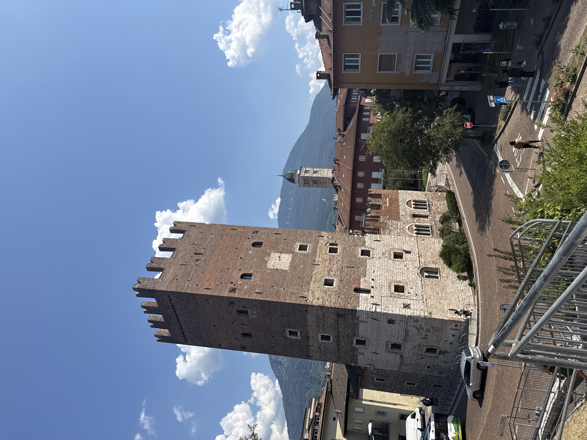 The image depicts a view of Torre Aquila, a historic medieval tower located in the city of Trento, Italy. The tower, constructed of red brick with stone accents at its base, stands prominently against a bright blue sky with scattered white clouds. Surrounding the tower are several multi-story buildings with terracotta tiled roofs, characteristic of Italian architecture. In the background, a mountain range is visible, partially obscured by trees and buildings. A street with cobblestone paving curves around the base of the tower, with several cars and a bus visible. A crosswalk with people crossing is in the foreground, along with a stop sign. The overall scene suggests a sunny day in an urban environment with historical significance.