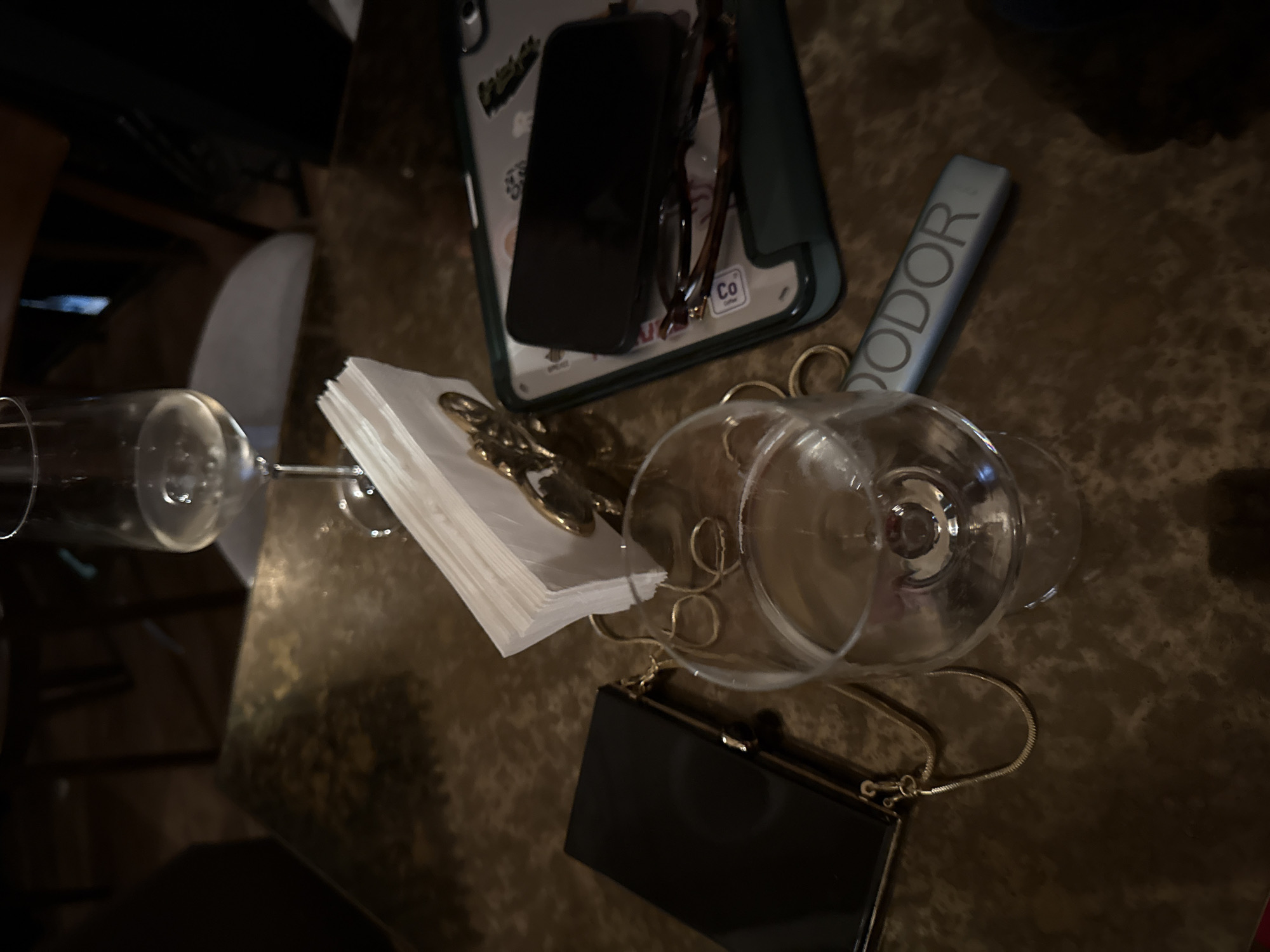 The image depicts a dimly lit, textured surface, possibly a table, with a disorganized arrangement of personal items. These include a cell phone lying atop a phone case, a pair of glasses, a small stack of napkins with a gold-colored object on top, a silver-colored tube labeled "ODOR," an empty wine glass lying on its side, and a small black clutch purse with a chain strap. The setting is not explicitly identifiable as Yerevan, Armenia, and no people, actions, or interactions are depicted. The environment is limited to the close-up view of the tabletop and associated objects.