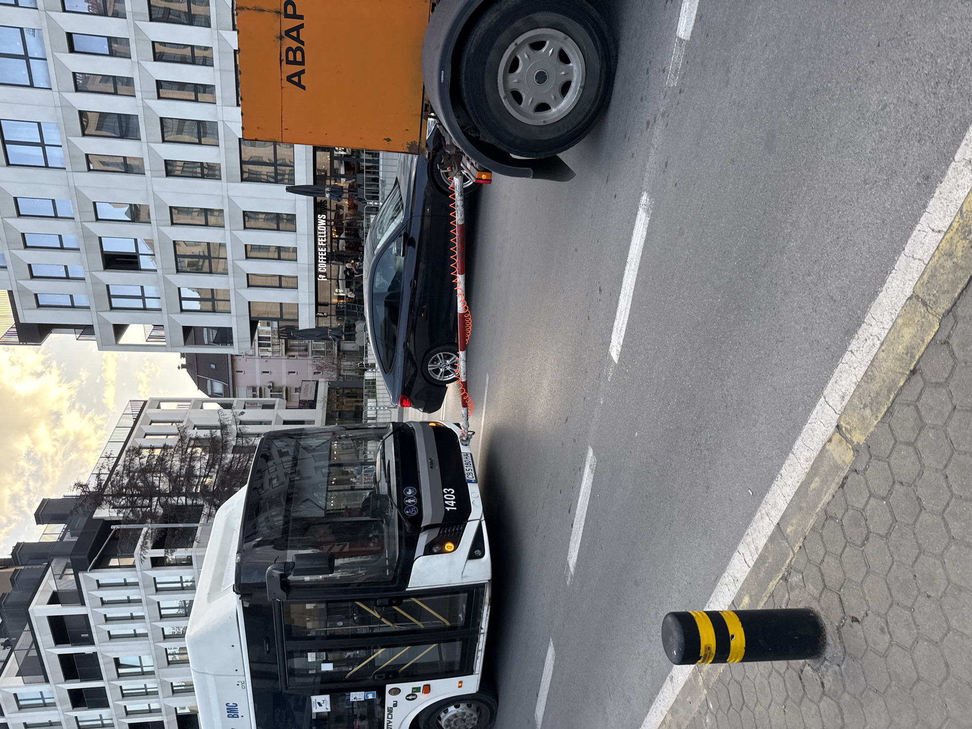 The image shows a street scene in Sofia, Bulgaria, featuring a BMC city bus numbered 1403 with license plate CB5180HA, parked on the left side of the frame. A black BMW car is partially obscured by an orange recovery vehicle with "ABAP" printed on its side. The BMW is being towed. Behind the recovery vehicle and car is a building with a restaurant named "Coffee Fellows". The location appears to be an urban area with modern architecture. A black and yellow bollard is visible on the right side of the frame, marking the edge of the road. There are no visible pedestrians or other vehicles in the foreground. The sky is visible in the background. The bus appears to be positioned on a designated lane marked by white lines on the asphalt.