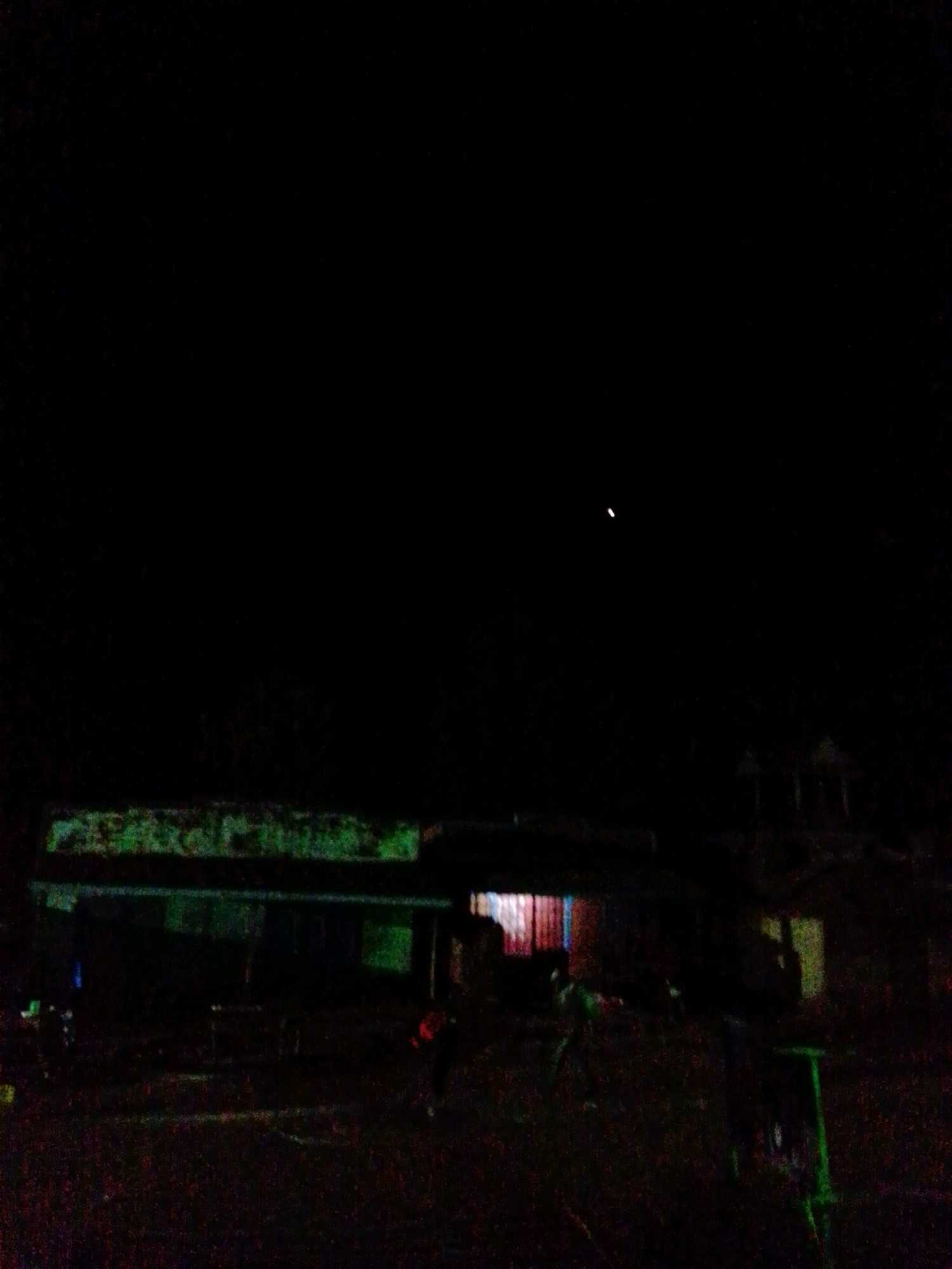 The image depicts a nighttime scene in Borabu, Chitago Ward, Kenya. In the foreground, a dimly lit, long, rectangular building with a green, possibly weathered, facade is visible. The building appears to have several sections or rooms with windows, some of which are illuminated from within, showing red and yellow hues. The ground in front of the building is dark and appears to be unpaved.
Several figures are present in the scene. To the left of the building, there are two individuals partially obscured by darkness, with one appearing to be wearing a blue garment. In the center, near the illuminated windows, a figure is seen wearing a green shirt, interacting with what looks like a bicycle or a similar object. Further to the right, another person in a red top is visible. In the far right of the image, a bright green structure, possibly a bench or a play structure, is present. The sky above is very dark, with a single bright, small object, possibly a distant light or a celestial body, visible in the upper middle section of the frame. The overall ambiance is that of a rural or semi-urban area at night, with minimal artificial lighting contributing to the low visibility.