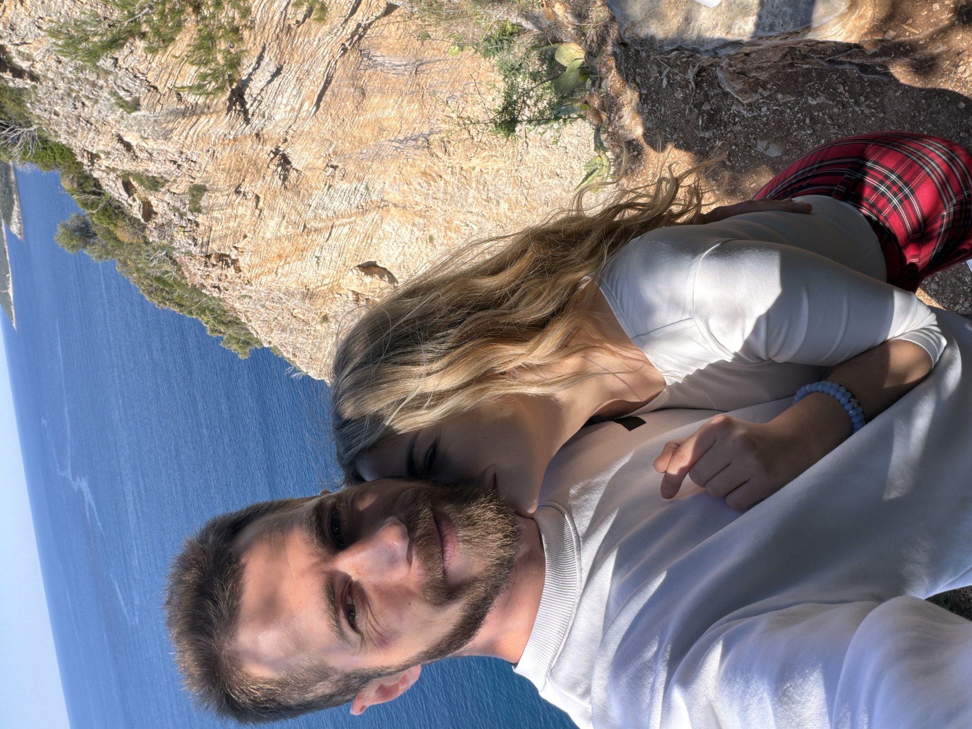 A couple is posing for a selfie on a cliff overlooking the Adriatic Sea in Budva, Montenegro. The man has short brown hair, a beard, and is wearing a white sweatshirt. The woman has long blonde hair and is wearing a white long-sleeved shirt and a red plaid skirt. She is leaning close to the man, her face near his. The background includes a rocky cliffside with vegetation and the clear blue sea. The image is taken from a slightly upward angle.