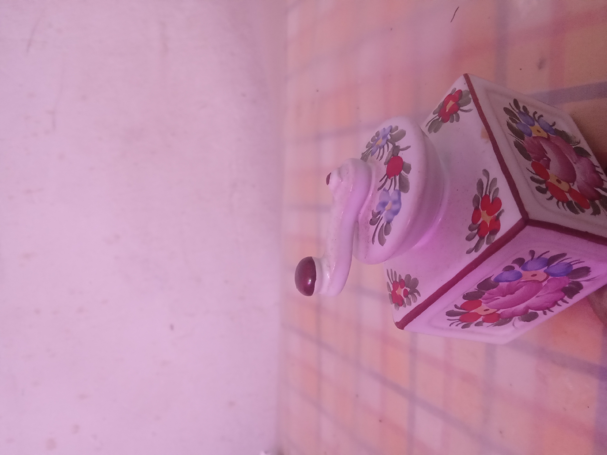 The image displays a decorative, miniature hand grinder, constructed from glazed ceramic or porcelain, exhibiting a primary white/off-white color scheme. Its square base is adorned with stylized floral motifs on each visible side, featuring broad strokes of red, magenta, blue, and green. A thin reddish-brown border frames the top edge of the base. A rounded top element, also white with smaller floral accents (blue and red flowers with green leaves), supports a curved handle terminating in a small, dark maroon spherical knob.
The object is positioned slightly off-center to the right, on a flat surface patterned with a grid of alternating light orange/peach and off-white squares, likely a tablecloth or a tiled surface. Small, dark particulate matter is visible on this surface. The background consists of a plain, light-colored wall, showing minor textural imperfections and faint dark specks. The entire scene is illuminated by a strong, pervasive pinkish-purple light source, casting a monochromatic tint across all visible elements and subtly altering their inherent colors. There are no individuals present, nor are any actions or interactions depicted. The precise location in the city of Unknown, Unknown, cannot be determined from the visual evidence.