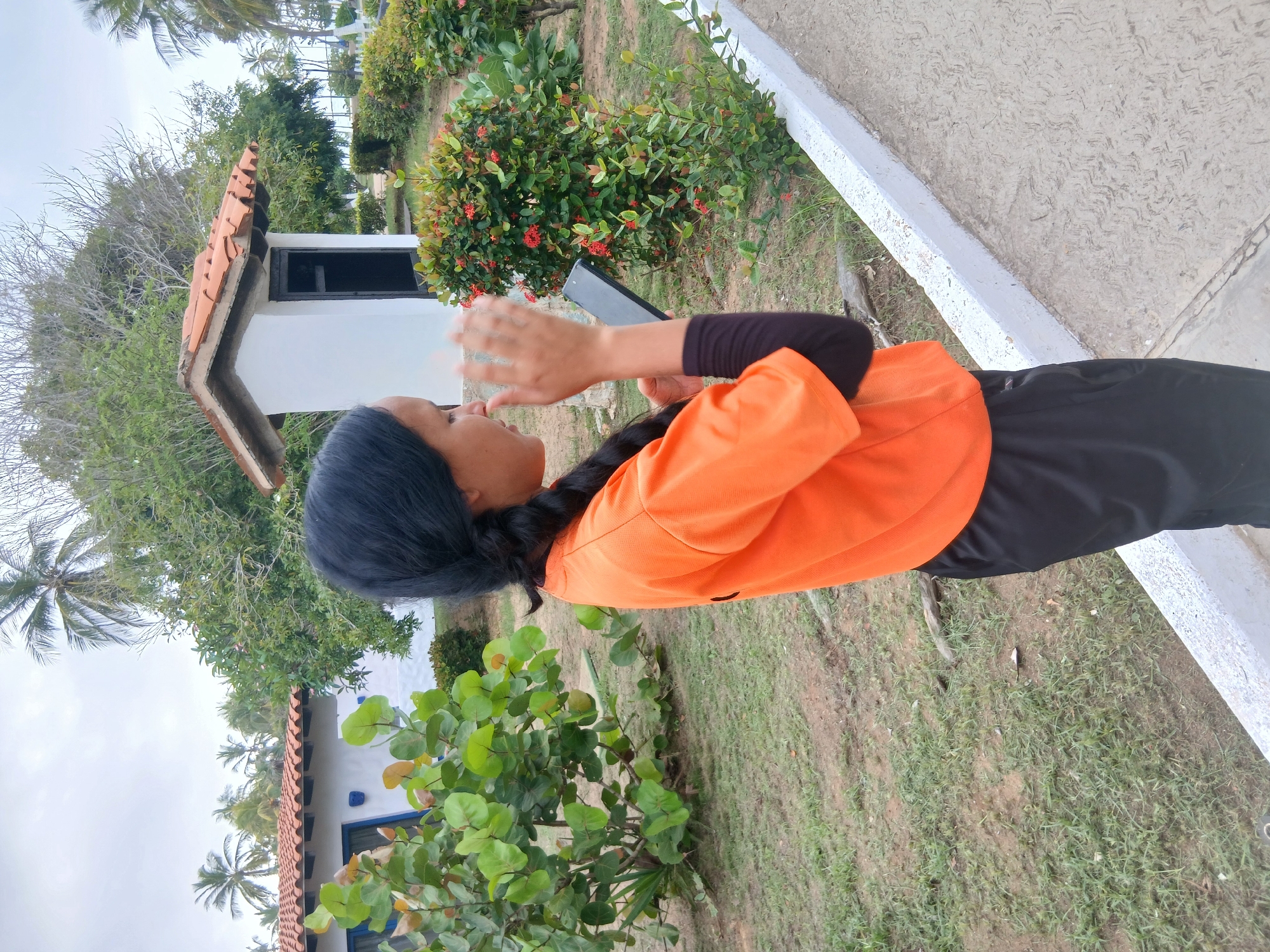 In Playa El Agua, Venezuela, a young woman with braided dark hair stands outdoors, holding a black smartphone in her right hand. She wears an orange short-sleeved shirt over a black long-sleeved undershirt and black athletic pants. Her left hand is raised, palm facing outward, in a gesture of speaking or gesturing. She is looking down at the phone, her expression neutral. The background features lush green foliage, including a large bush with red flowers, a white building with a terracotta tiled roof, and palm trees. The ground is covered with patchy grass and a dirt path. The overall setting appears to be a tropical park or resort area.