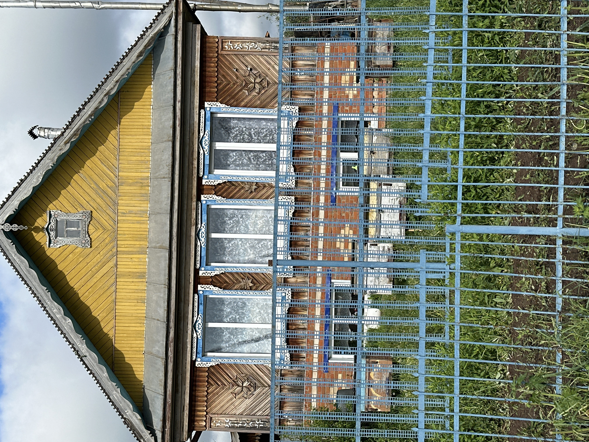 The image captures a view through a blue metal fence, revealing a portion of a wooden house. The house is located in Среднее Кечево, Russia. The house features a yellow wooden facade and a roof with metal gutters. There are three visible windows with decorative blue trim and patterned glass. The wooden portion of the house has intricate carvings. Behind the wooden part of the house, there is a section constructed of red bricks. The ground visible through the fence contains dirt and green vegetation.