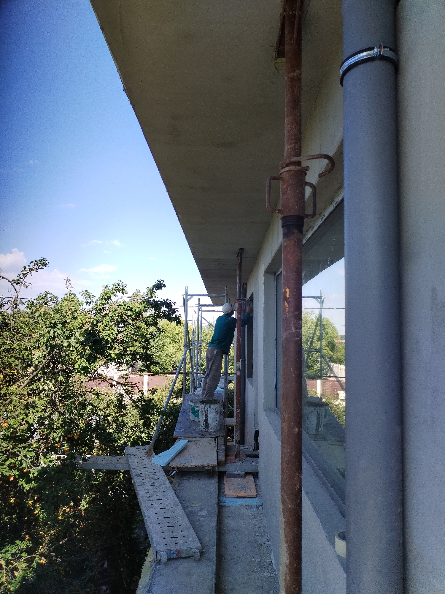 A lone construction worker, wearing a teal long-sleeved shirt, grey pants, and a white hard hat, is on scaffolding outside a building, applying sealant around a window frame. The worker is positioned to the left of a large grey pipe and a rusted metal support pole. The scene is set against a backdrop of lush green trees and a clear blue sky with a few scattered clouds. The building appears to be under construction or renovation, with visible scaffolding and building materials. The image is taken from a high angle, looking down towards the trees and rooftops visible behind the scaffolding.