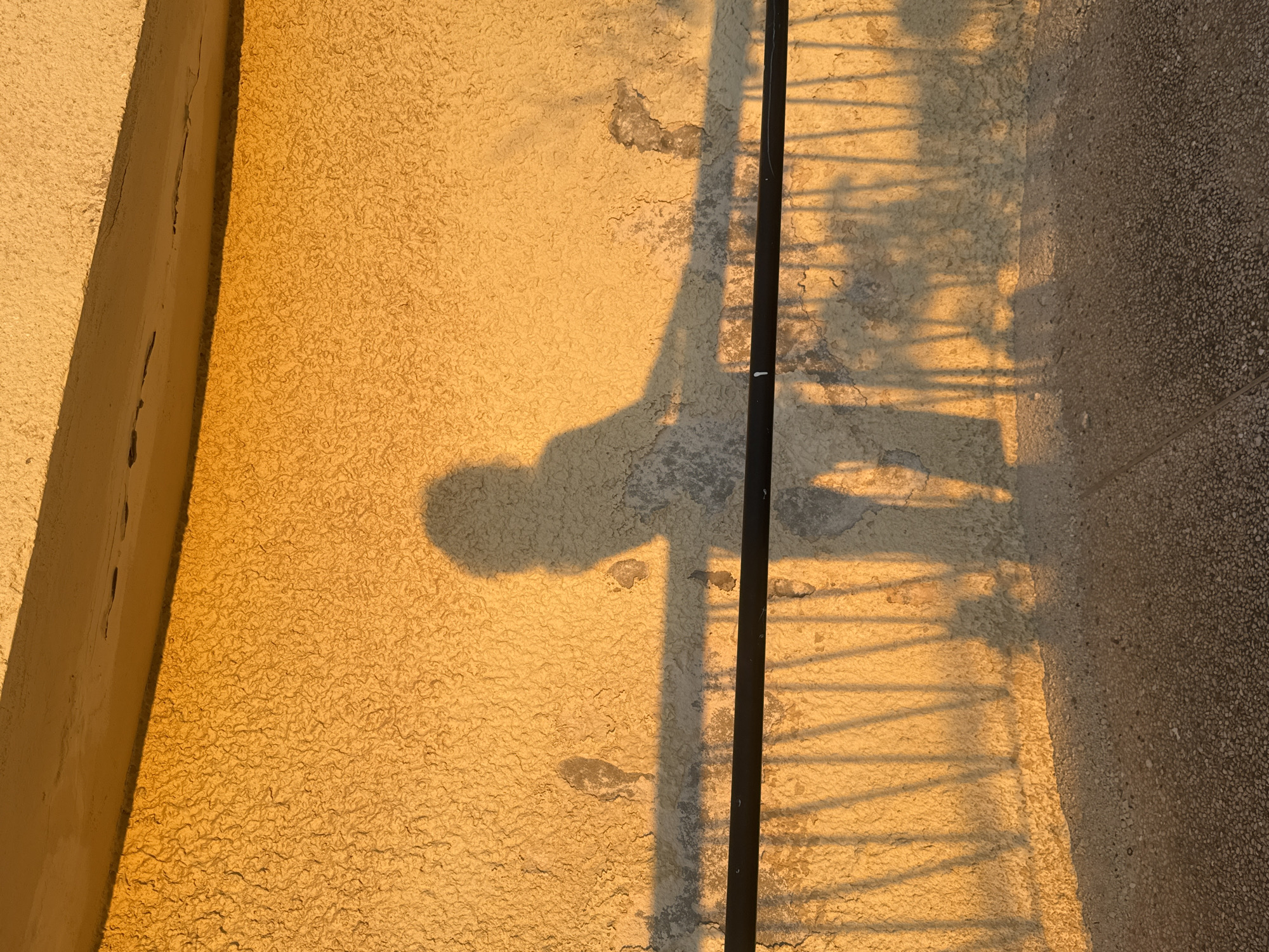 The image depicts a shadow of a person leaning on a railing against a textured, rough, light yellow wall. The shadow is cast by the warm, golden light of what appears to be sunset or sunrise, creating a stark contrast between the illuminated wall and the darker shadow. The railing, a dark horizontal bar with vertical supports, casts its own shadow pattern on the wall, intersecting with the person's shadow. The wall itself shows some signs of wear and tear, with patches of lighter material and subtle discoloration. Below the wall is a textured ground surface, likely a pathway or pavement, also illuminated by the same warm light. The scene suggests an outdoor urban setting, possibly a balcony, walkway, or public space in Ramat Gan, Israel, given the context of the location. The overall impression is one of quiet contemplation or a moment of pause, captured by the interplay of light and shadow.