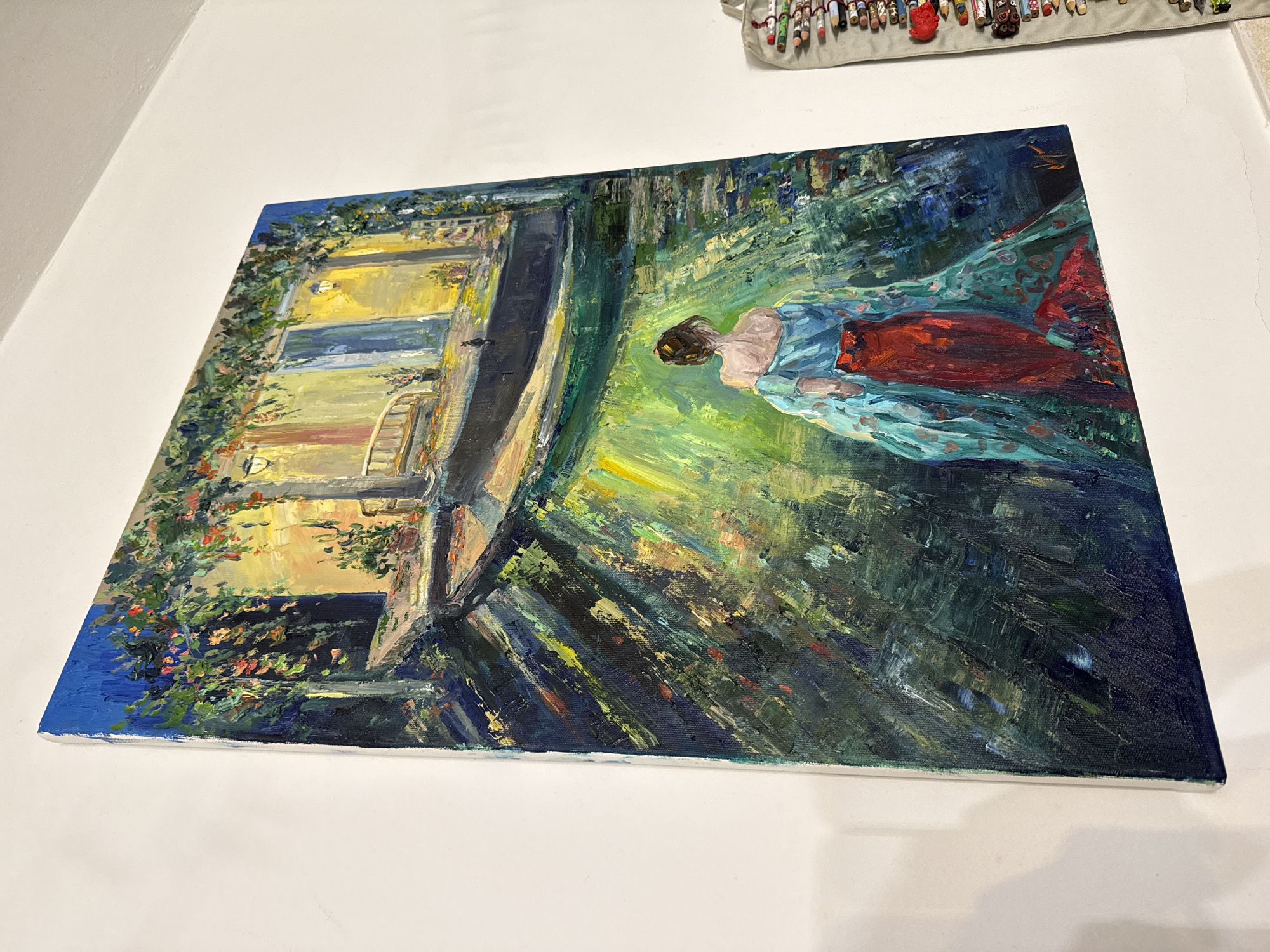 An oil painting depicts a woman from behind, wearing a flowing blue and red gown, walking away from the viewer down a pathway. The pathway is illuminated by dappled sunlight filtering through lush green foliage overhead, casting strong shadows. In the background, a building with yellow walls, blue doors, and glowing lanterns suggests a European setting. The style is impressionistic, with visible brushstrokes adding texture and depth. The painting is hung on a white wall, and to the right, a hanging organizer filled with art supplies is partially visible. There is no indication in the image that this scene is specifically located in Herceg Novi, Montenegro.