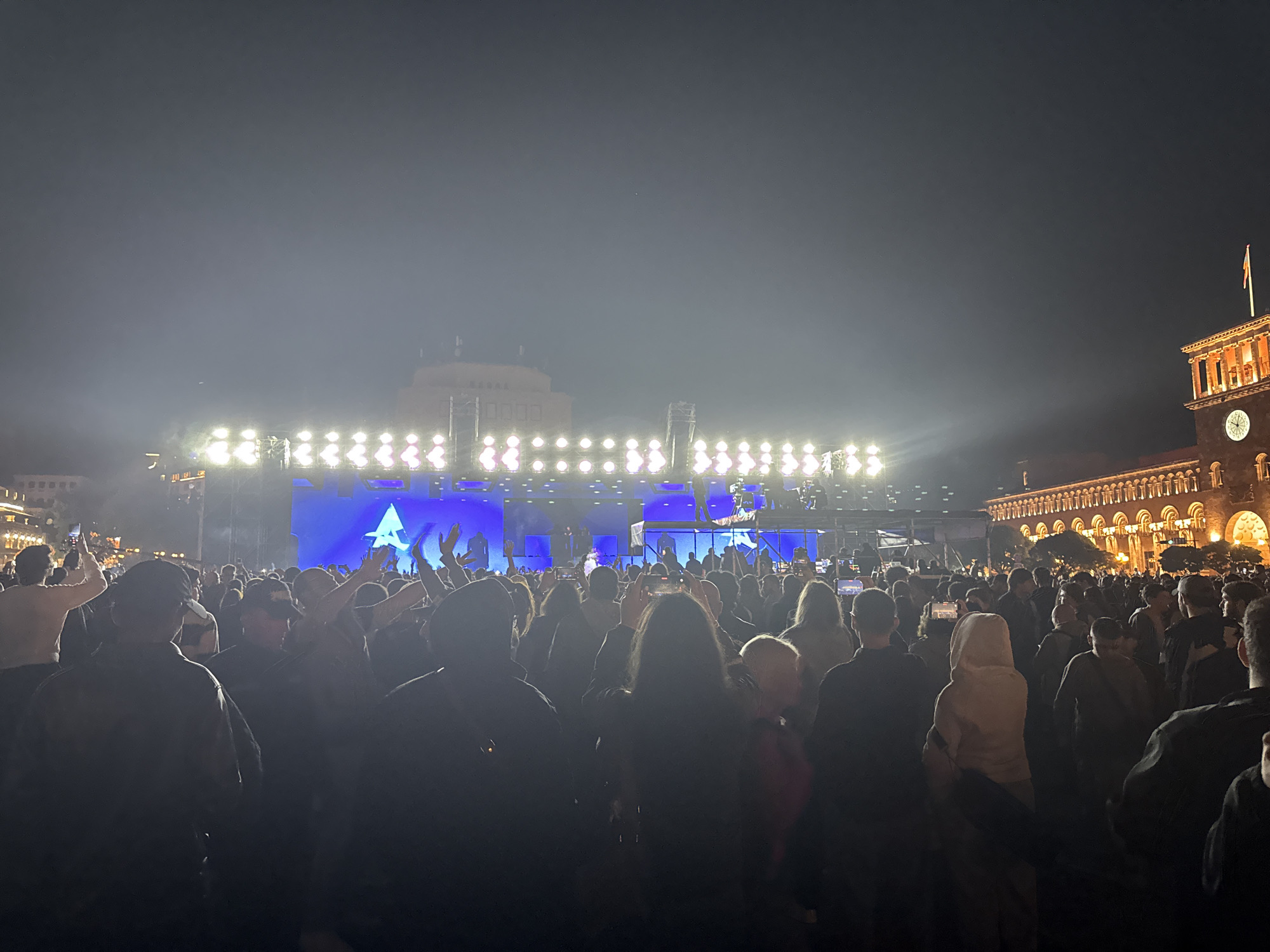 A large crowd of people gathers at night in a city square for an outdoor event. A brightly lit stage stands at the forefront, with a large screen displaying a blue background and a symbol resembling the letter "A." The crowd, mostly silhouetted against the bright stage lights, is dense and stretches back into the distance. Many individuals are holding up their hands, suggesting they are enjoying the performance. A building with a clock tower is visible on the right side of the frame, and another building is discernible in the background behind the stage.