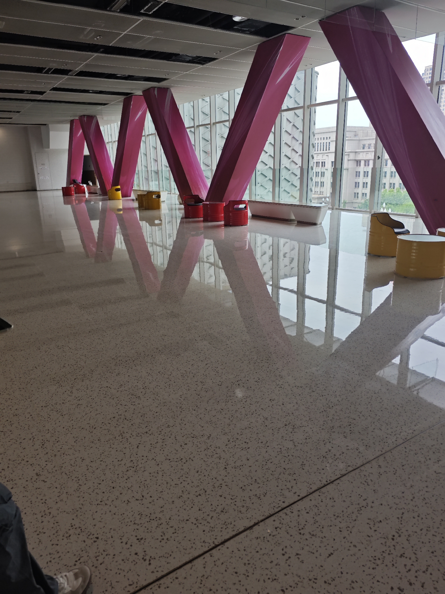The image shows an indoor space, possibly a lobby or waiting area, with several bright pink, angled support columns. The shiny floor reflects the columns and the light coming through the wall of windows. The window offers a view of a building in the distance. Several red and yellow, barrel-shaped objects are placed along the floor near the base of the columns, and a white bench is located in the middle of the room. The photograph's position is at ground level in front of the objects. A person is wearing denim clothing in the bottom left corner of the picture. Based on the location provided, the architectural style and the surrounding environment, this is most likely an indoor public space in the Liangjiang New Area in Chongqing, China.