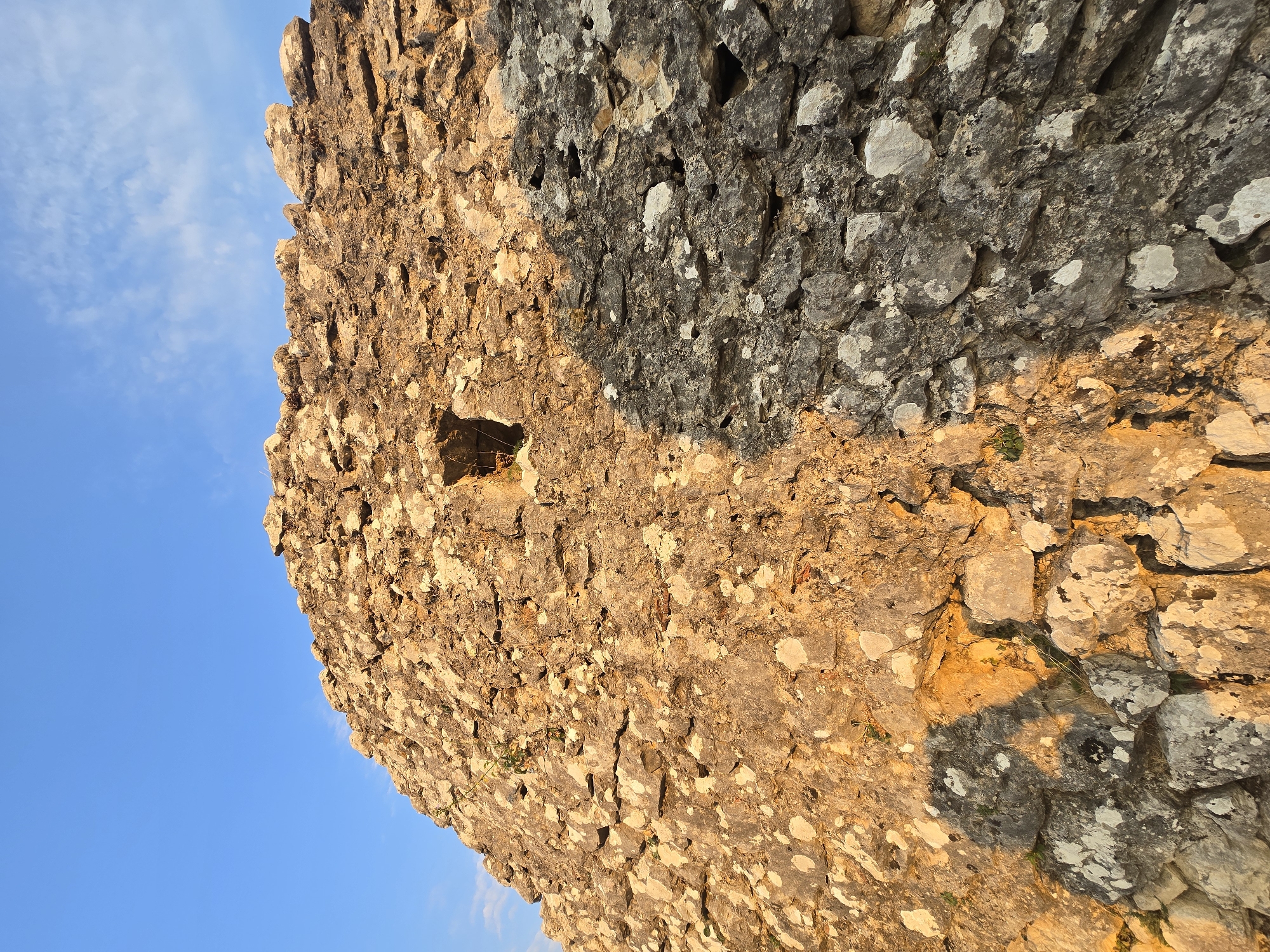 The image captures a close-up view of an ancient stone wall, likely part of the ruins of Medun Fortress in Medun, Montenegro. The wall is constructed from irregular, weathered stones of varying sizes, some lighter in color and others darker, with patches of lichen. Sunlight casts a warm glow on the left side of the wall, highlighting its texture and creating shadows. A small, dark opening, possibly a window or crenel, is visible in the upper central portion of the illuminated section. The sky above is a clear, vibrant blue with a few wispy white clouds. The overall impression is one of historical decay and natural beauty.