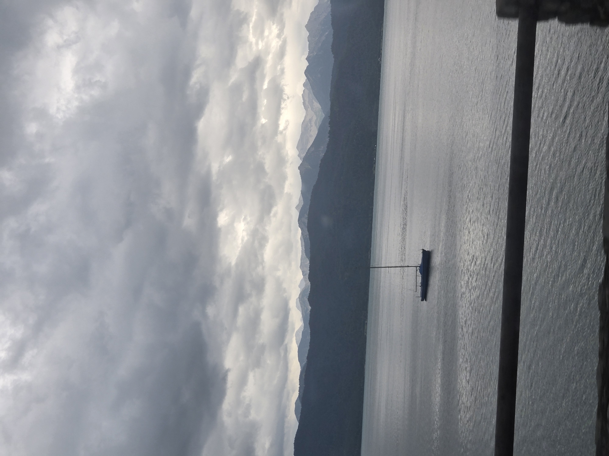 A sailboat is anchored on a calm lake in front of a tree-covered mountain range under a cloudy sky. The lake is dark blue and reflects the overcast sky. The mountains in the background are dark green and covered in trees. The sky is filled with gray clouds. A wooden railing runs along the bottom of the image. The scene is a serene and peaceful landscape.