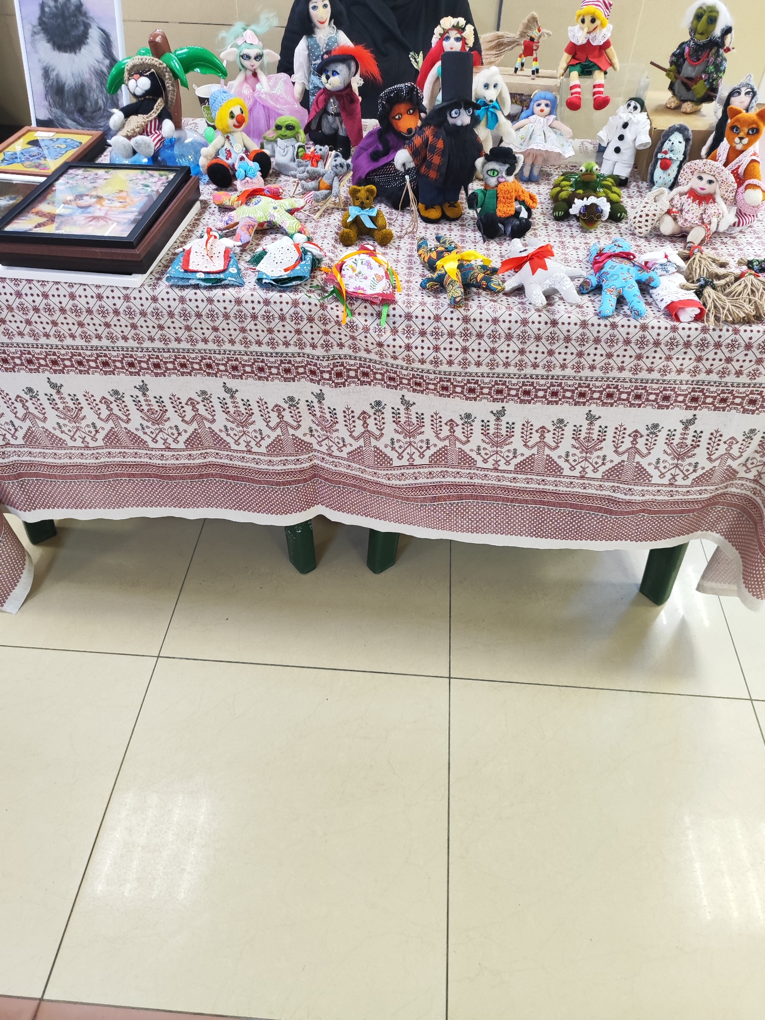 The image shows a table covered with a tablecloth, displaying numerous handmade dolls and crafts. The tablecloth is white with a red patterned border featuring geometric and stylized floral designs. The crafts include various dolls, some depicting animals, fairytale characters, and figures in costumes. Some framed pictures are also visible on the table. The table is positioned on a tiled floor, with the tiles appearing to be a light beige color.
The location, Fryazino, Russia, can only be inferred from external data not included in this image. Therefore, any mention of the location is speculative. The scene is a craft display, possibly at a fair or market.