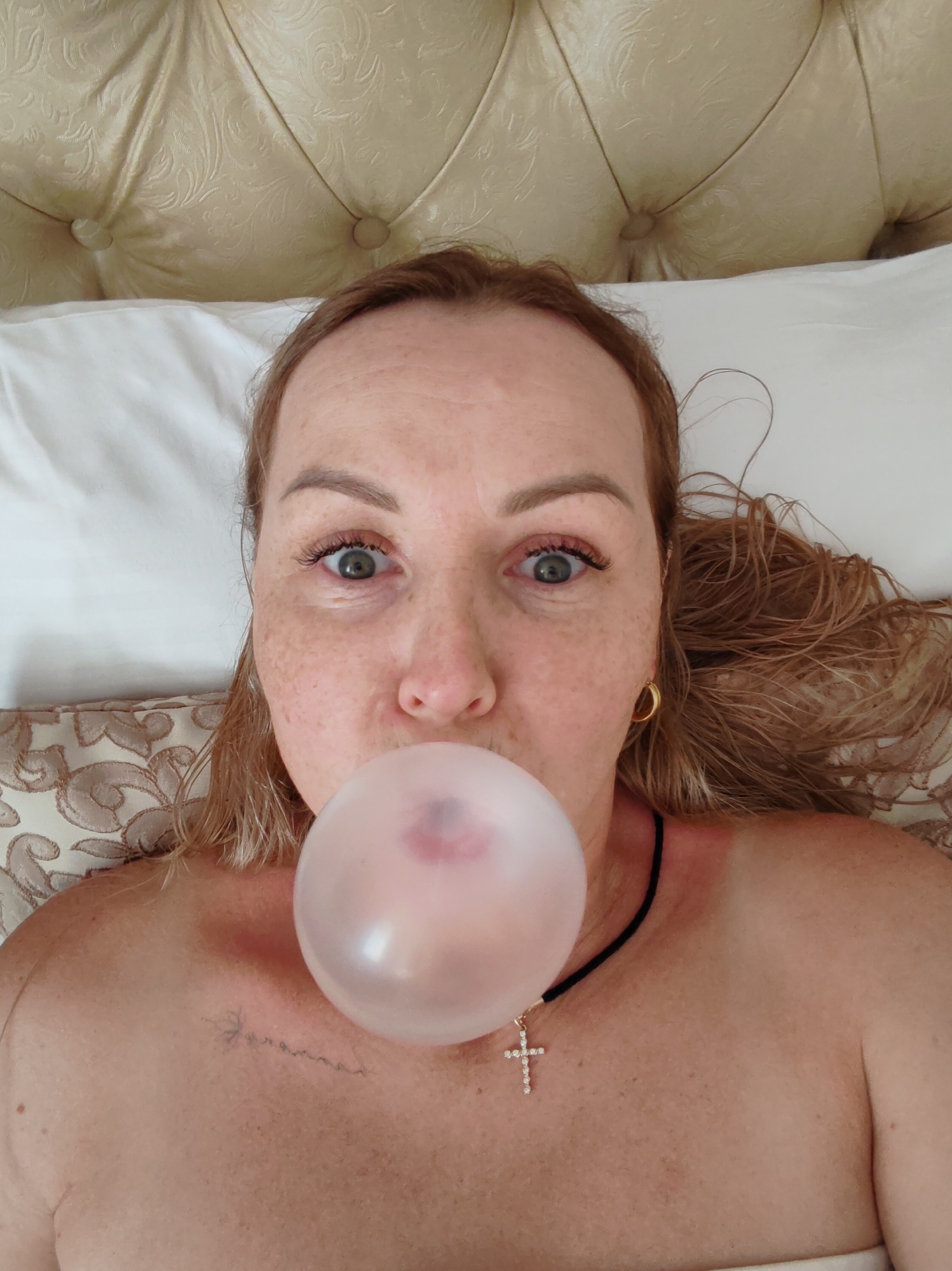 A woman with light skin, freckles, and light brown hair lies on a bed with a light-colored headboard and white pillows. She is blowing a pink bubble gum bubble. She is wearing a black choker necklace with a silver cross pendant and gold hoop earrings. The woman has a tattoo on her upper left chest. The location is not specified.
