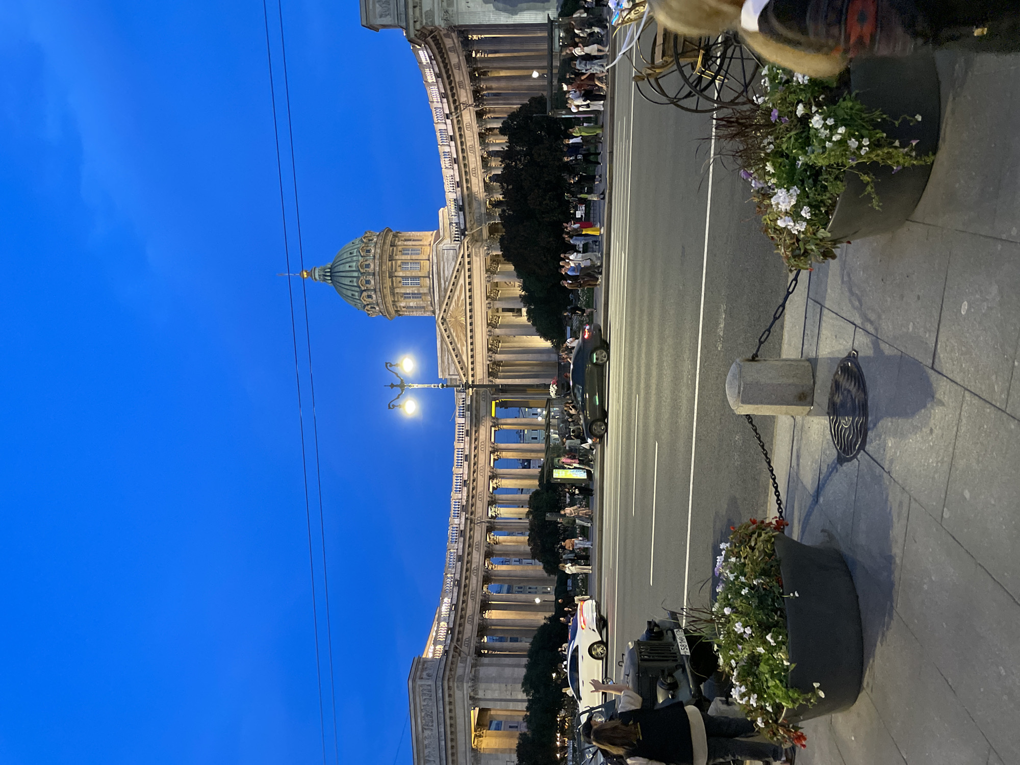 A wide shot captures the Kazan Cathedral in St. Petersburg at dusk. The iconic blue sky is visible above the grand semicircular colonnade of the cathedral. In the foreground, a street is bustling with activity, featuring a white car, a dark sedan, and several pedestrians. Two large planters filled with white and purple flowers are positioned on the sidewalk, adding a touch of nature to the urban landscape. The overall scene evokes a sense of a vibrant city at twilight, with a blend of architectural grandeur and everyday life.