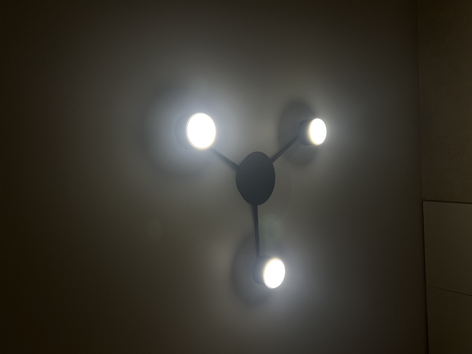 A modern black ceiling light fixture with three illuminated bulbs hangs from a dark ceiling. The fixture has a central hub from which three arms extend, each terminating in a bulb. The bulbs emit a bright, white light, casting shadows on the ceiling. The lower portion of the image shows a portion of a white cabinet. This scene is indoors, and the specific location in Sheptytskyi, Ukraine is not discernible from the image.