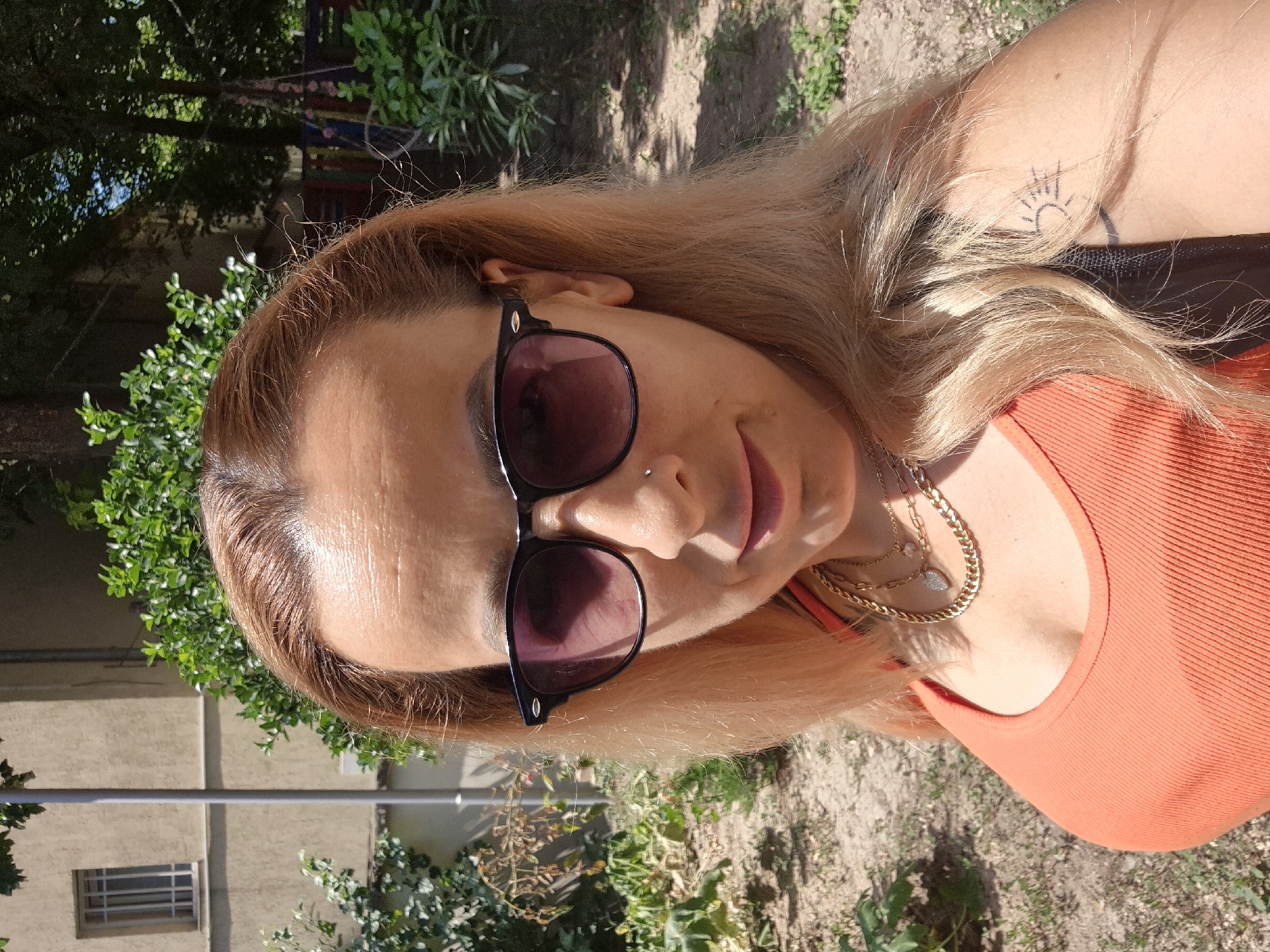 A woman is taking a selfie outdoors in front of a building. She has blonde hair and is wearing sunglasses and an orange tank top. She is also wearing a gold necklace with a heart pendant. There is a tattoo of a sun on her right arm. The background shows a building, trees, and bushes. The lighting suggests it is daytime.