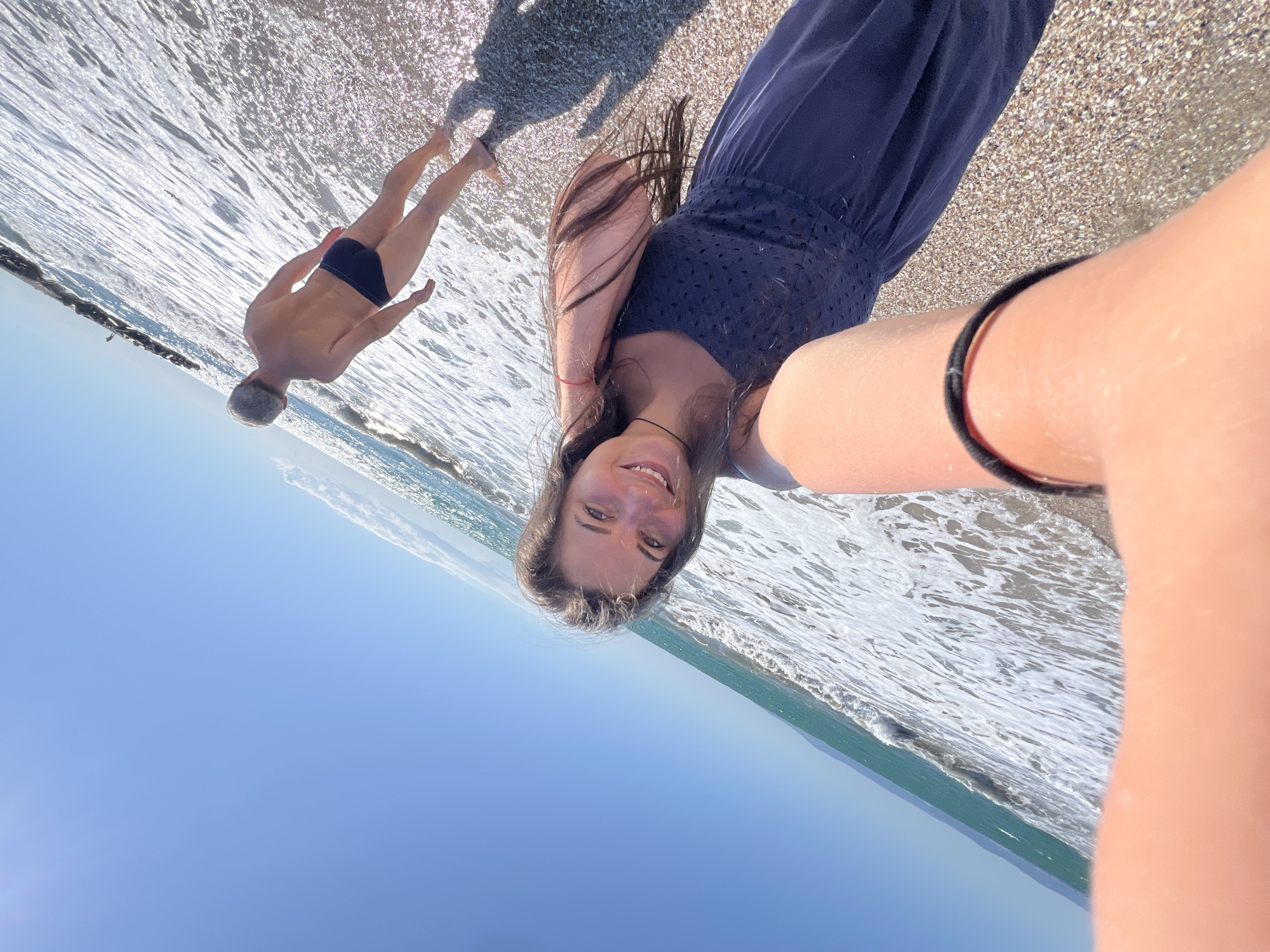 In the city of Pomorie, Bulgaria, a young woman with long, dark hair and a blue dress smiles as she takes a selfie on a beach. The sun is bright, casting shadows on the pebbled shore and sparkling on the shallow waves. In the background, a man with grey hair, wearing dark blue swimming trunks, walks away from the camera towards the sea. The ocean extends to the horizon under a clear blue sky, with a breakwater made of rocks visible in the distance. The scene captures a moment of leisure and enjoyment on a sunny day by the Black Sea.