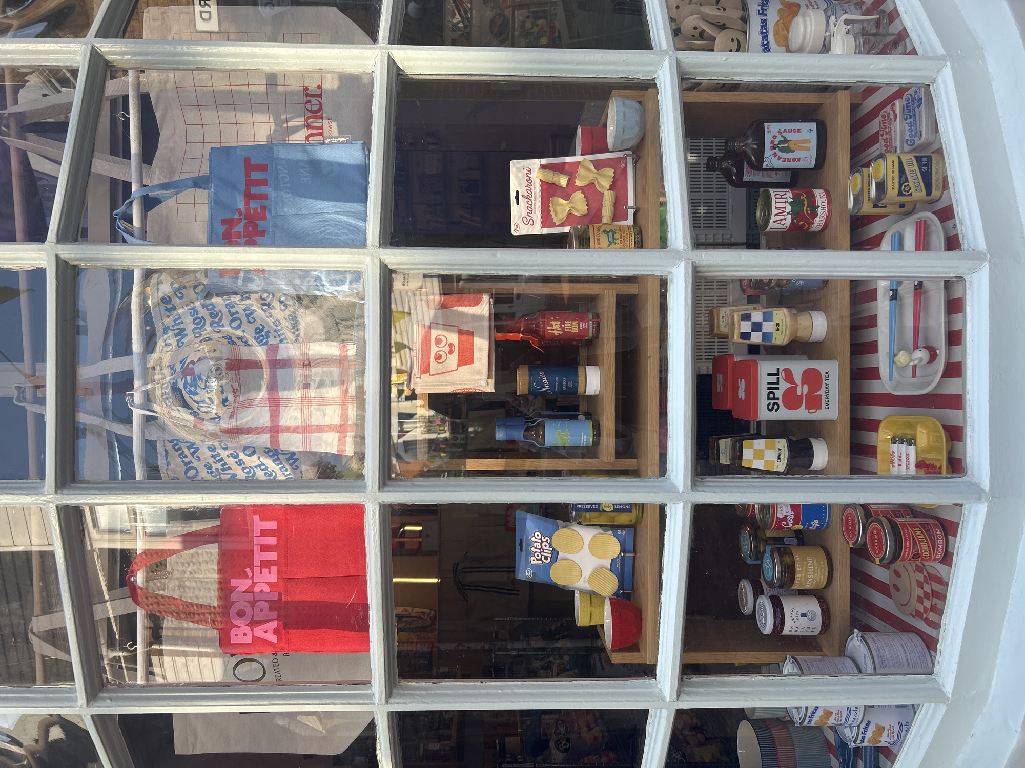 The image shows a shop window display in Brighton, United Kingdom. The window is divided into multiple panes by white mullions. The display features a variety of food-related items and merchandise.
In the top row of panes, there are three tote bags. The left pane shows a red tote bag with the words "BON APPETIT" in pink lettering. The middle pane has a red and white checkered tote bag. The right pane displays a light blue tote bag with "BON APPETIT" in red lettering and a grid pattern.
The middle row of panes showcases more items. The left pane has a display of "Potato Clips" potato snacks, two bowls (one red, one yellow), and a jar of preserved lemons. The center pane features several bottles and jars, including a bottle labeled "Hot Fire Bow", a bottle of "Naiise Mayonnaise", and a bottle of what appears to be soy sauce. The right pane has a package of "Snackaroni" pasta, a jar of "Rodeo Beef Jerky", and two small bowls (one red, one light blue).
The bottom row of panes contains an assortment of packaged goods and kitchenware. The left pane shows stacked bowls or containers and cans labeled "Tostadas Fritas". The center pane displays several jars, including one labeled "GOCHUJANG BOM BOM", and a yellow tray with chopsticks and a small figurine. The right pane features various bottles and jars, including a can labeled "AMIRI SHAKSHOUKA", a bottle of "KOREAN BBQ SAUCE", and smaller bottles. There is also a can labeled "SPILL EVERYDAY TEA".
The overall impression is of a quirky and eclectic shop selling artisanal food products and novelty items. The sunlight casts reflections on the glass, and the items are arranged on wooden shelves and a striped background.